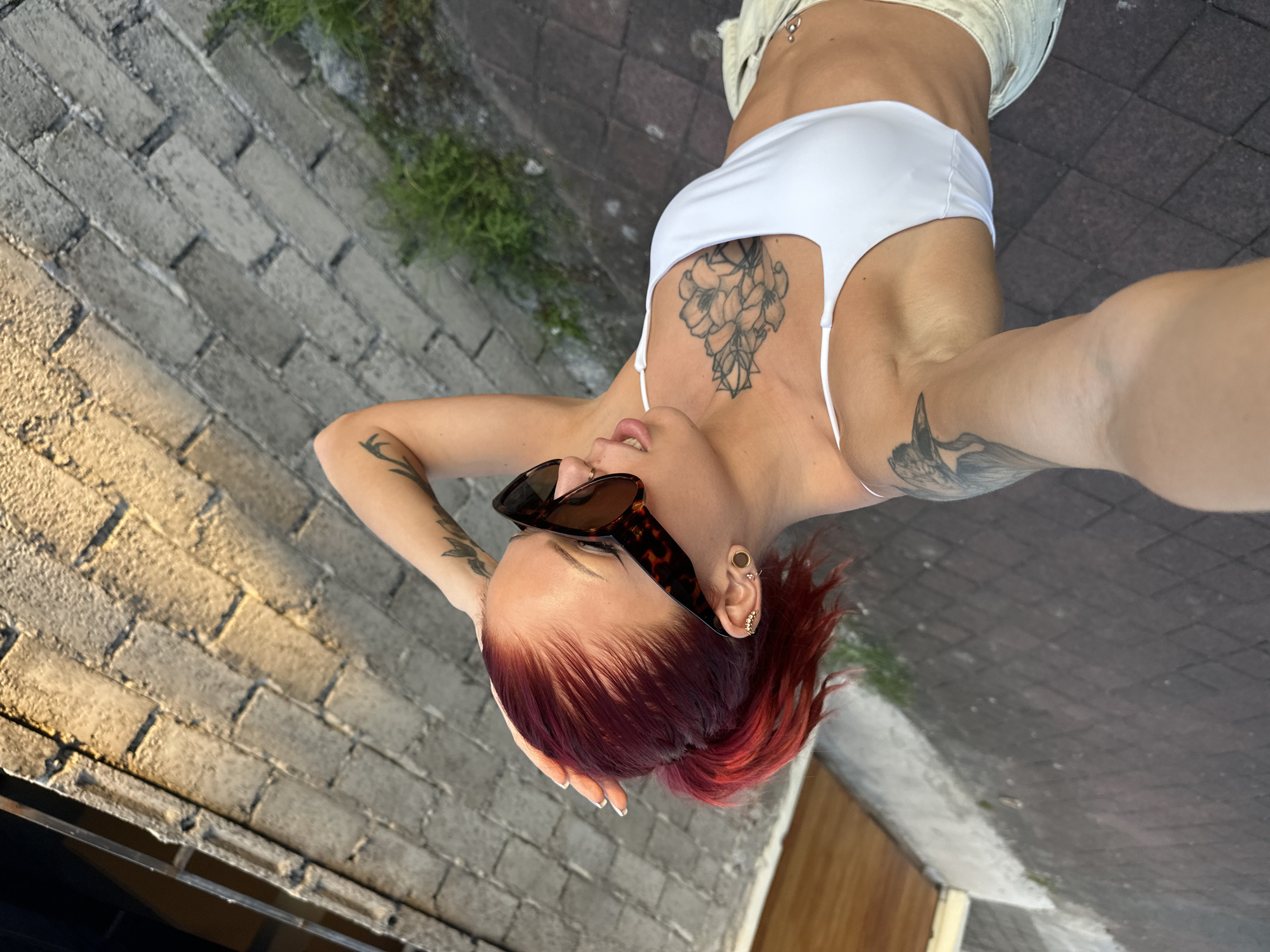 A woman stands on a brick path in what appears to be an outdoor setting, holding the camera and taking a selfie. She is dressed in a white top and light-colored shorts. She wears sunglasses and has reddish-brown hair and multiple visible tattoos on her arms and chest. The location could potentially be North Kuta, Indonesia, however, the image does not have any other items that indicate this location. The background features brick structures and some vegetation.