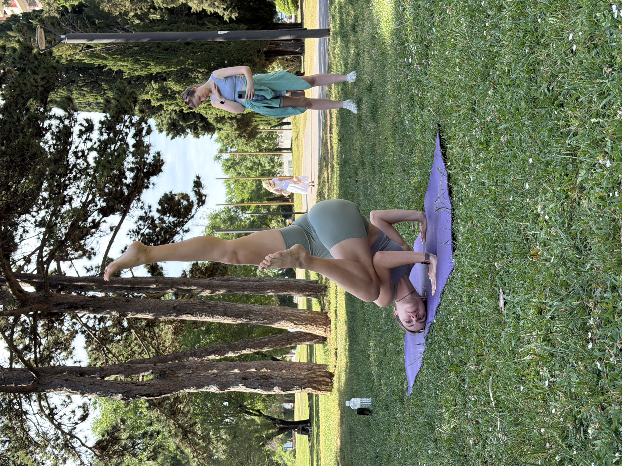 A sideways view of a grassy park scene shows a woman in green yoga attire, in the tripod headstand pose on a purple yoga mat. Her legs are extended straight up into the air, her head is down, and her hands are supporting her head. A water bottle and a phone are next to her on the grass. In the background, another woman, in a blue top and green skirt, is holding a phone. The location may be in or near Budva, Montenegro. Trees are in the background on the left.