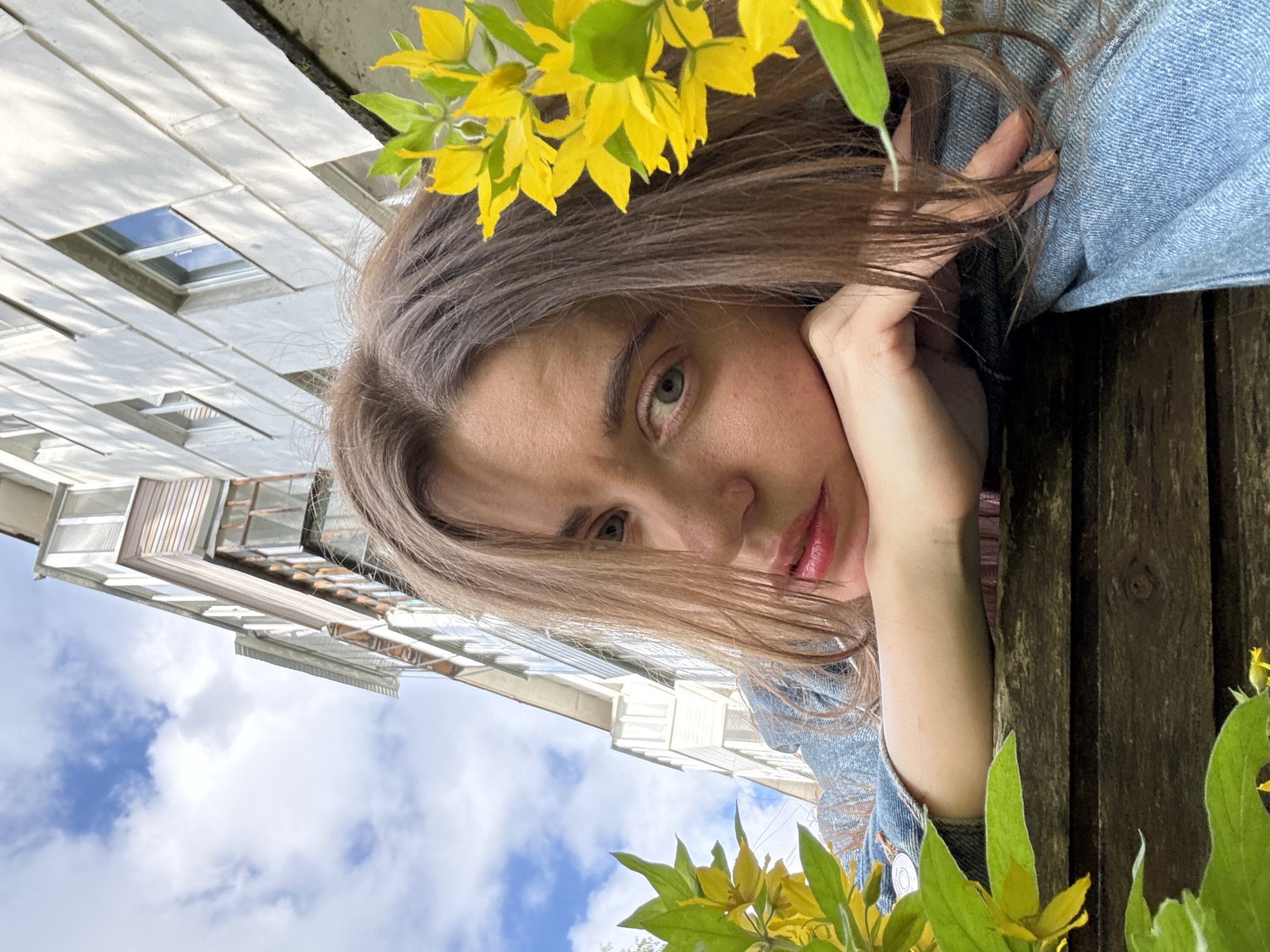 A young woman with long, brown hair rests her chin on her arm, looking towards the camera. She wears a denim jacket and is positioned outdoors, with a building and sky in the background. Yellow flowers and green leaves are visible in the foreground, partially obscuring the woman. The perspective is a low-angle shot, looking upwards.