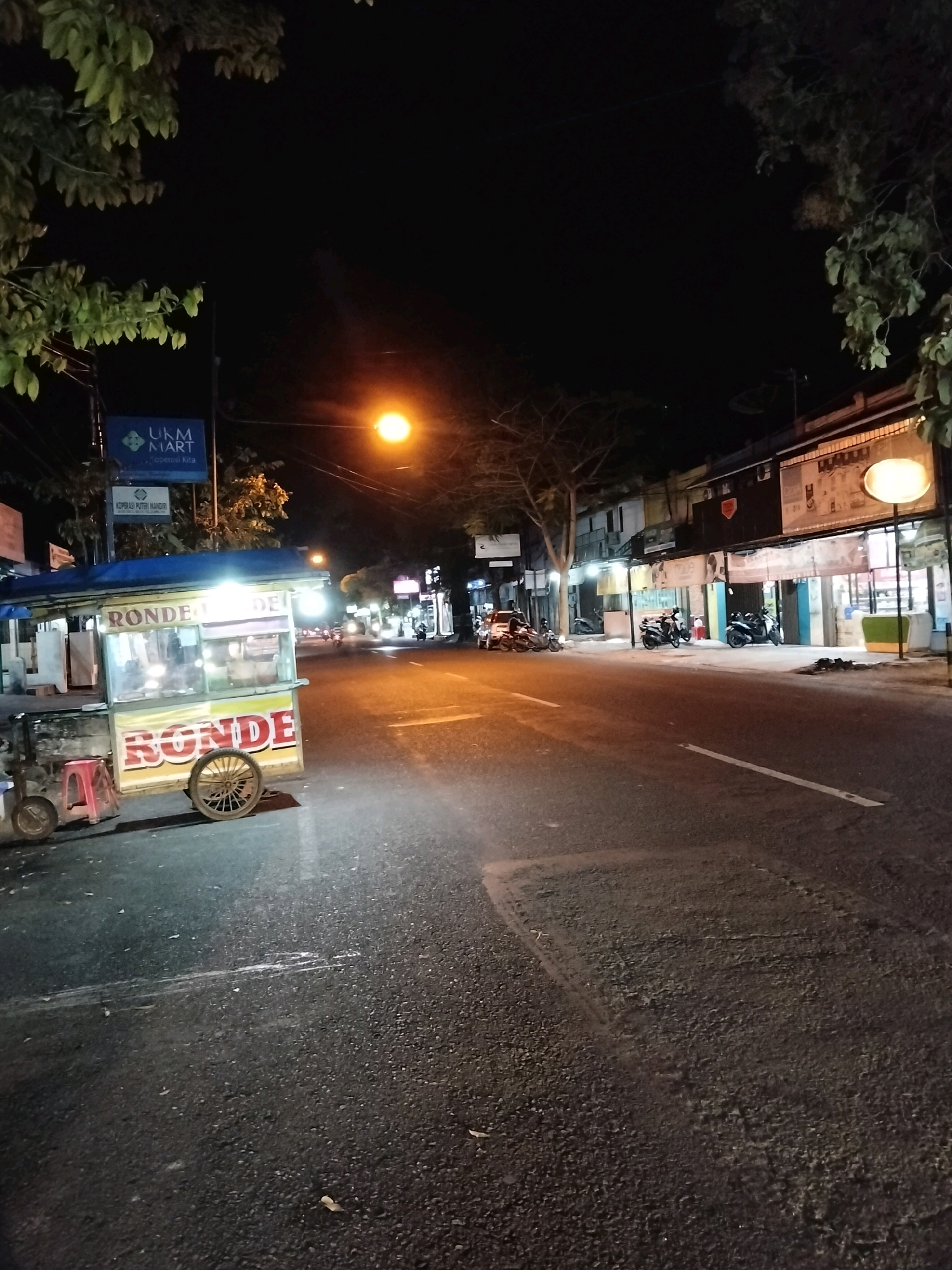 The image depicts a street scene at night in Purworejo, Indonesia. On the left side of the road, a food cart with the word "RONDE" prominently displayed is parked. The cart is illuminated by overhead lights and appears to be open for business. A red stool is placed next to it. Further down the street, there are several motorcycles parked and a few individuals can be seen walking or riding. Buildings line the right side of the street, some of which have illuminated storefronts. A street lamp is visible in the mid-ground, casting a warm glow on the road. Trees with foliage are present on both sides of the street, their leaves illuminated by the artificial lights. The sky is dark, indicating it is nighttime. The road surface is asphalt and appears wet in some areas, reflecting the light. A traffic sign reading "UKM MART" is visible on a pole behind the food cart. The overall impression is that of a typical urban street scene at night with commercial activity.