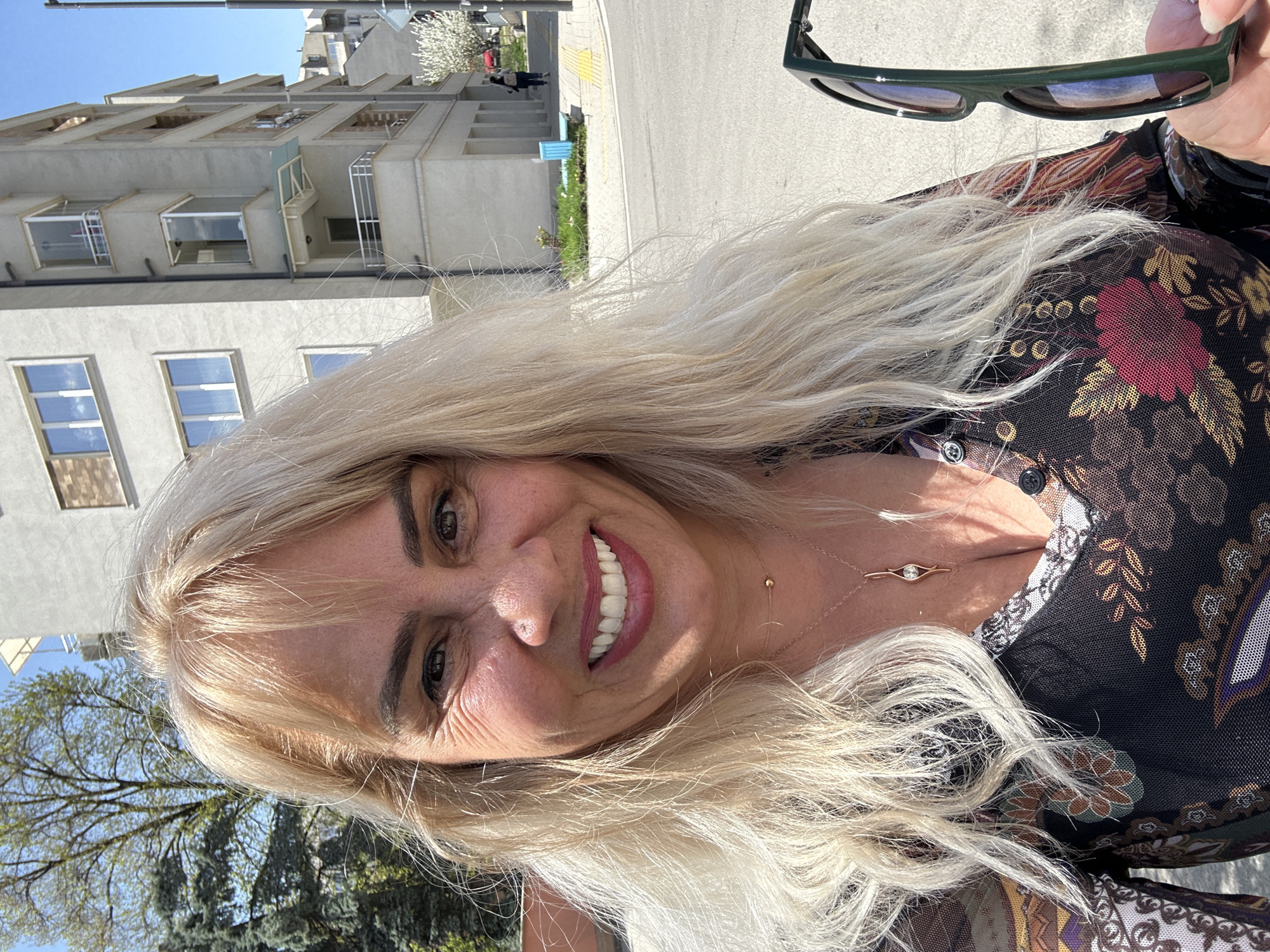 The image captures a selfie of a smiling woman outdoors on a bright, sunny day, likely taken in Sofia, Bulgaria. The woman, who appears to be in her 40s or 50s, has long, flowing blonde hair that cascades around her shoulders. She looks directly at the camera with a wide, cheerful smile, revealing bright white teeth. Visible wrinkles are present around her dark eyes, and she has a small nose piercing. She is dressed in a dark-colored top or dress adorned with a rich floral and patterned design in shades of maroon, gold, and brown. Around her neck, a delicate gold chain holds a small eye-shaped pendant and a tiny gold bead. In her right hand, held towards the upper right of the frame, are dark green sunglasses.
The background behind her suggests an urban or suburban environment. A clear blue sky is visible above several light-colored, multi-story buildings featuring numerous windows and some small balcony-like structures. To the woman's left, green trees, both evergreen and deciduous with burgeoning spring leaves, are visible. To her right, a paved street or sidewalk with distinct yellow markings stretches into the distance, where a lone figure can be seen walking and a light blue object is present. The strong, natural sunlight indicates it is daytime, casting clear shadows. No visible text appears in the image. The overall impression is one of happiness and contentment.