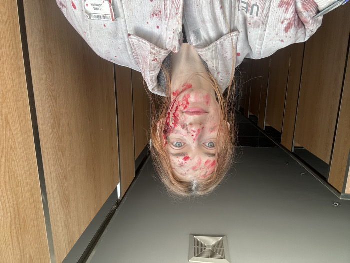 A female individual, appearing to be Caucasian with reddish-blonde hair and blue eyes, is depicted in an indoor public restroom setting within Huangdao District, China. She is looking directly at the camera with a neutral expression, potentially a slight smile. Her face, particularly the left side around the temple and ear, as well as her nose and right cheek, and portions of her hair, are covered in extensive red liquid and dark smudges consistent with blood. She is wearing a light-grey or white collared garment, resembling a jacket or coverall, which is heavily splattered with numerous red stains also consistent with blood. A name tag on her left chest (viewer's right) reads "EMMA JOHNSON" with a barcode above it. A partial logo or text, "UEI," is visible on her right chest (viewer's left). The background consists of a series of light-wood laminate partition doors with vertical metal frames, typical of toilet stalls. A square ventilation fan is mounted centrally on the light-colored ceiling.