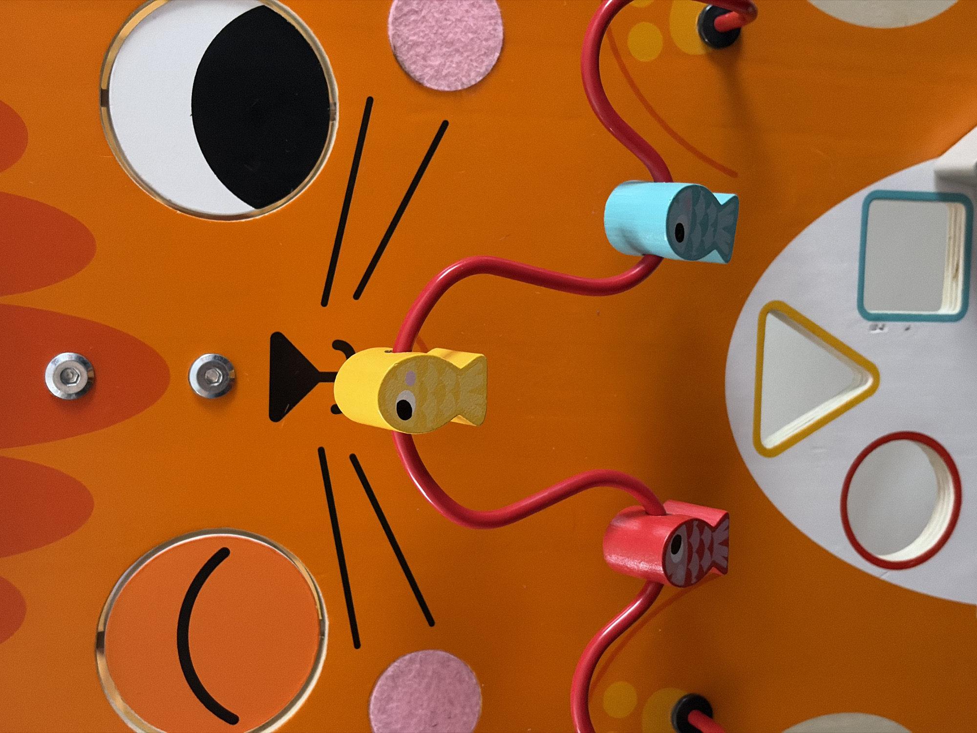An extreme close-up, overhead shot shows a section of a children's wooden activity toy. The toy is predominantly orange, designed to resemble a cat's face with large circular eyes, one with a black pupil and the other a closed eyelid with a black curved line. Black lines represent the cat's whiskers and a small black triangle forms its nose.
Two pink fuzzy circles are attached to the orange surface on either side of the face, likely representing cheeks or ears. A red wire bead maze snakes across the surface. Attached to this maze are three wooden animal figures, shaped like fish. One yellow fish hangs above the nose area, and a red and a blue fish are positioned lower down on either side, threaded onto the red wire.
Below the cat face are white cutouts in geometric shapes, including a circle with a red border and a triangle with a yellow border. A small blue-bordered square is also visible, which appears to be a small mirror. The overall impression is a colorful and engaging toy designed for young children. The location is Unknown, Unknown.