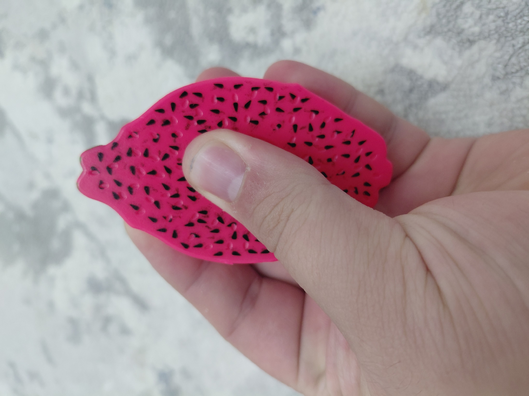 In Aktau, Kazakhstan, a hand holds a bright pink, stylized representation of dragon fruit, its surface dotted with black seeds. The object is shaped like a slice of dragon fruit, complete with the characteristic lobed edge. The background consists of a light-colored, textured surface, possibly a table or floor, with a mottled grey and white pattern. The lighting is even, suggesting an indoor or shaded outdoor environment. The image is a close-up, focusing on the interaction between the hand and the fruit replica.