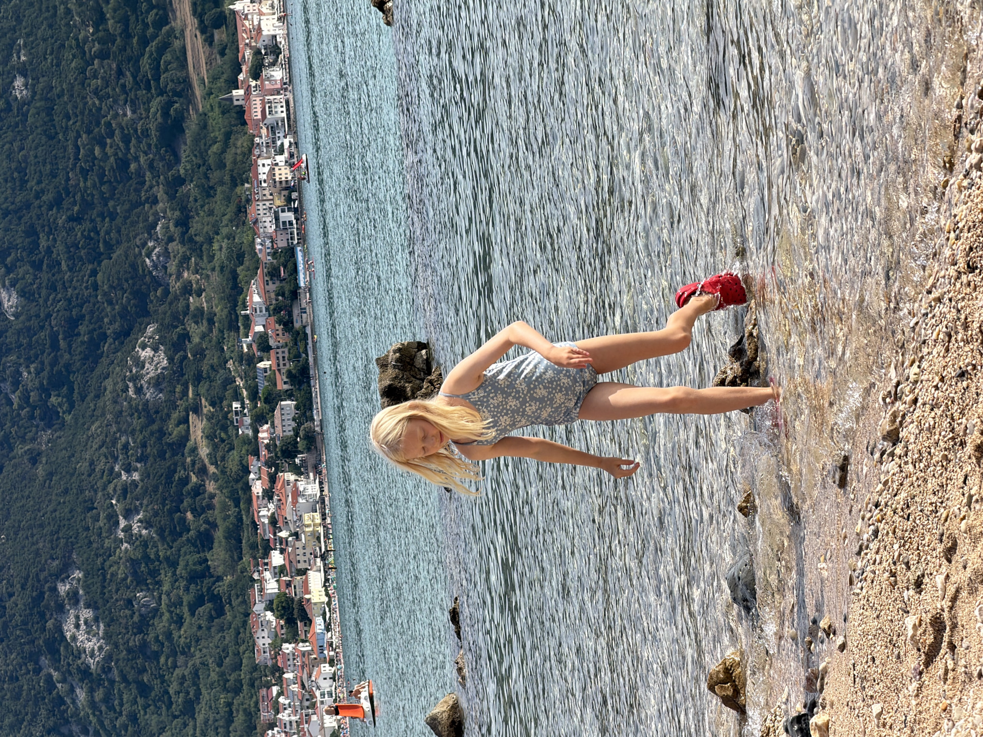 A young girl with blonde hair and a floral-patterned romper is standing on a pebbly beach with her feet in the shallow, clear water. She is wearing red shoes. The girl appears to be reaching her hand into the water. The background shows a densely forested hillside dotted with white buildings and red-tiled roofs, indicating a coastal town. The water is calm with small ripples, and the sun is bright, suggesting a sunny day. The scene evokes a sense of a peaceful summer vacation. The location is identified as Općina Baška, Croatia. There is no visible text in the image.