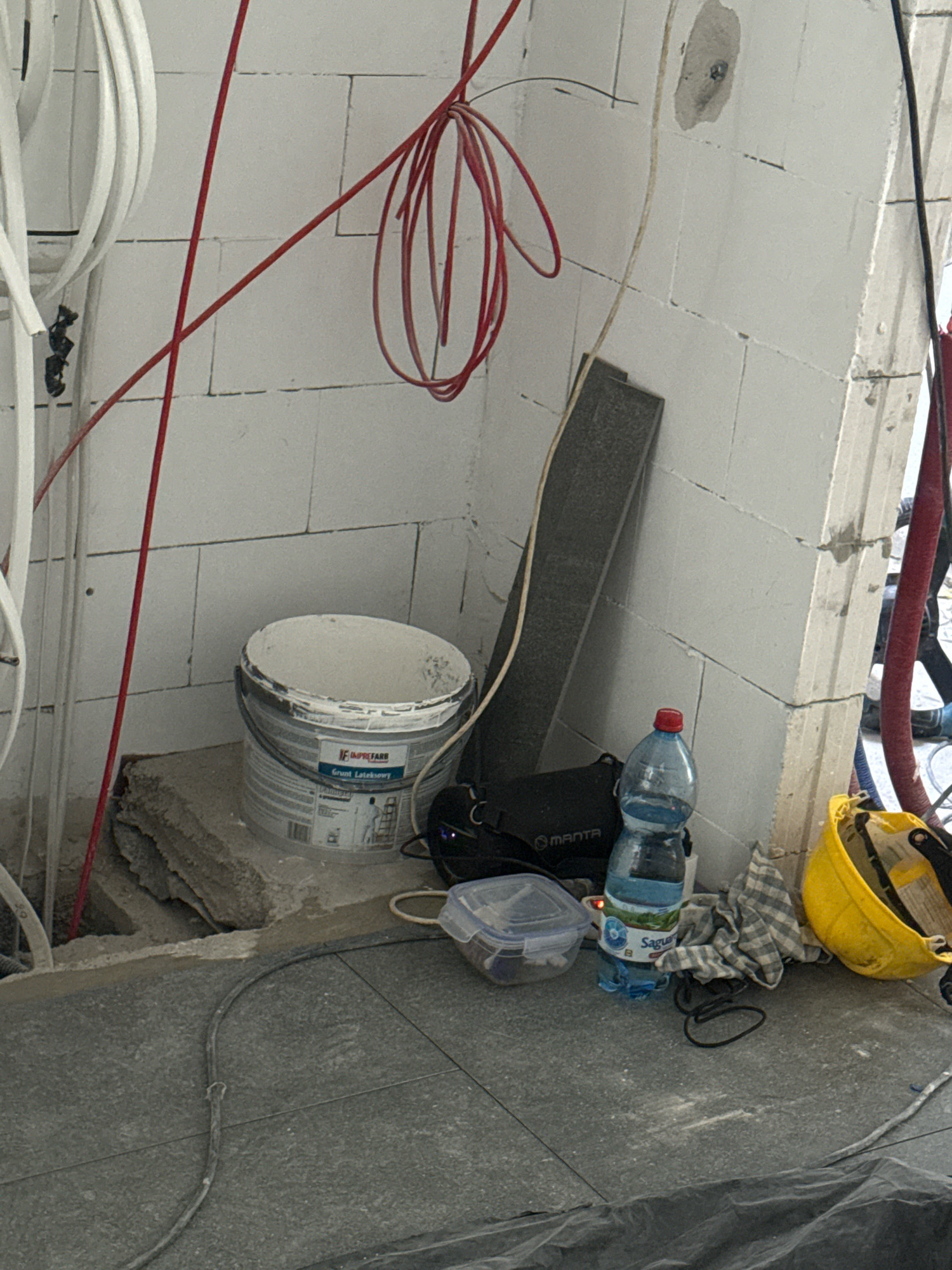 The image depicts an indoor construction site in Nowa Sól, Poland. The environment is characterized by unfinished white brick walls, exposed electrical wiring, and concrete floors. Several construction-related items are visible: a large white bucket labeled "IMPREFARF Grunt Lateksowy," a blue plastic water bottle, a black bag with the "MANTA" logo, a yellow hard hat, and various lengths of coiled red and white cables. A clear plastic container with contents is also present on the floor. The overall scene suggests an active construction or renovation project in progress.