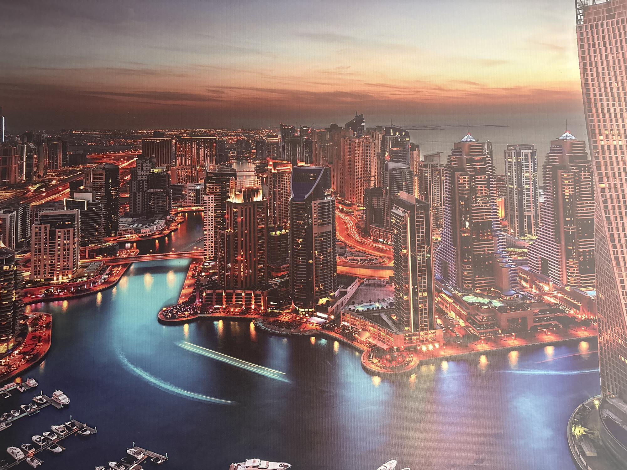 This is an aerial or high-vantage view of a modern, densely populated cityscape at dusk or early night. The scene features a vast expanse of high-rise skyscrapers, illuminated with warm artificial lights, stretching across the landscape. A prominent, winding canal or bay cuts through the urban area, with buildings lining its banks.
The sky above shows the remnants of a sunset, displaying warm orange, red, and purple hues near the horizon, gradually fading into darker tones overhead. The city's numerous lights, from buildings, streets, and bridges, reflect vividly on the dark, calm surface of the water, creating a mesmerizing glow. Streaks of light from moving vehicles are visible on the roads and bridges, indicative of a long-exposure photograph, highlighting the constant flow of traffic.
Along the waterfront, several marinas are visible, housing numerous boats and yachts docked in neat rows. The illuminated promenades and commercial establishments at the base of the buildings suggest a vibrant and active urban environment. The contemporary architectural style, the extensive waterfront development, and the overall grandeur of the urban planning strongly suggest a location such as Dubai, particularly the Dubai Marina area.
The image captures the dynamic energy and modern aesthetic of a major global city, emphasizing its architectural achievements and bustling nightlife. There is no visible text in the image.
