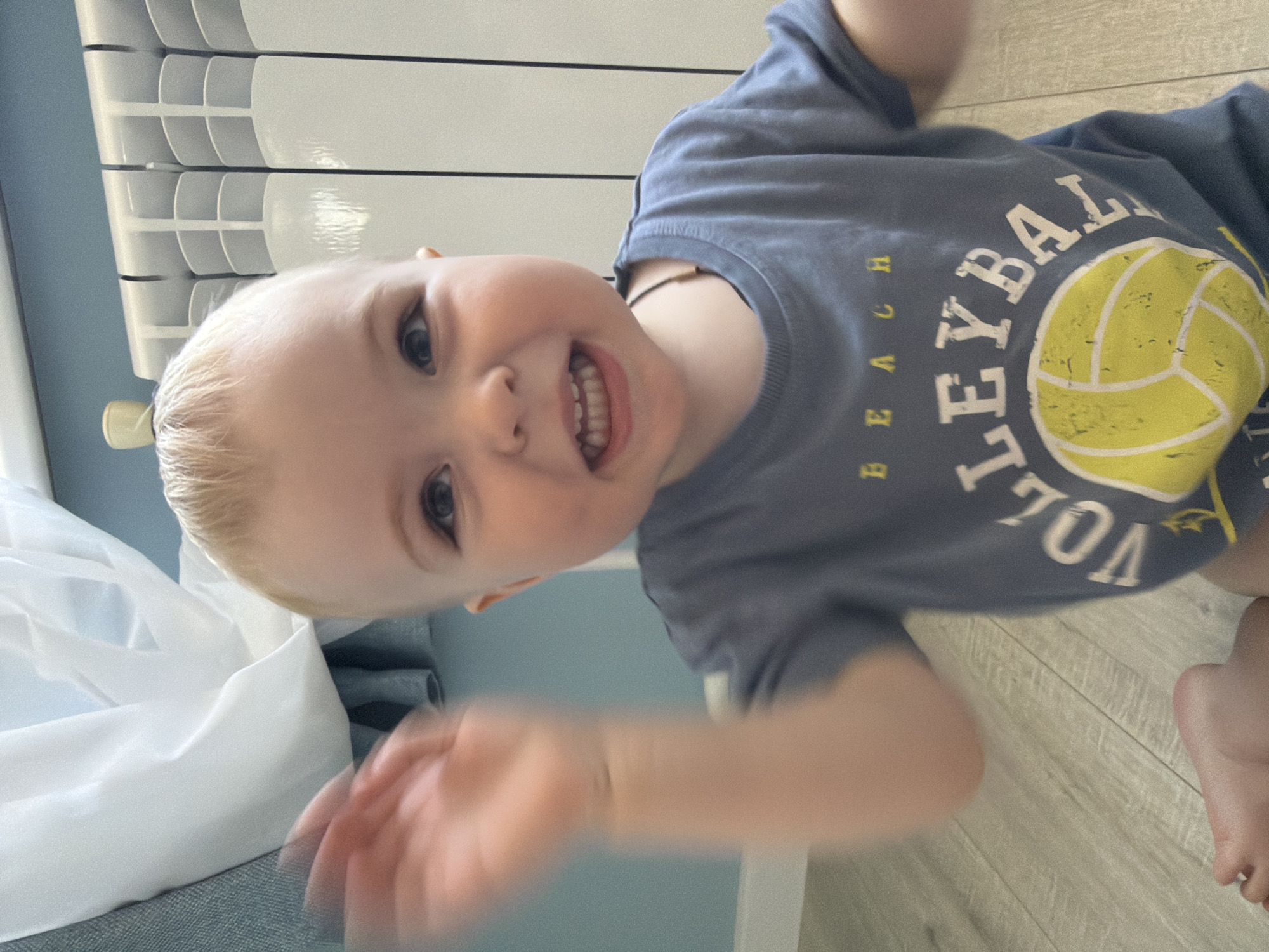 A close-up, slightly tilted shot depicts a young child with fair hair and a wide smile, wearing a blue t-shirt with a yellow volleyball design and the word "VOLLEYBALL" printed on it. The child's hand is blurred in the foreground, suggesting movement. Behind the child is a white radiator and a light blue surface, possibly a wall or piece of furniture. The floor appears to be a light wood. The location is likely indoors, but no specific landmarks or details point to Votkinsk, Russia.