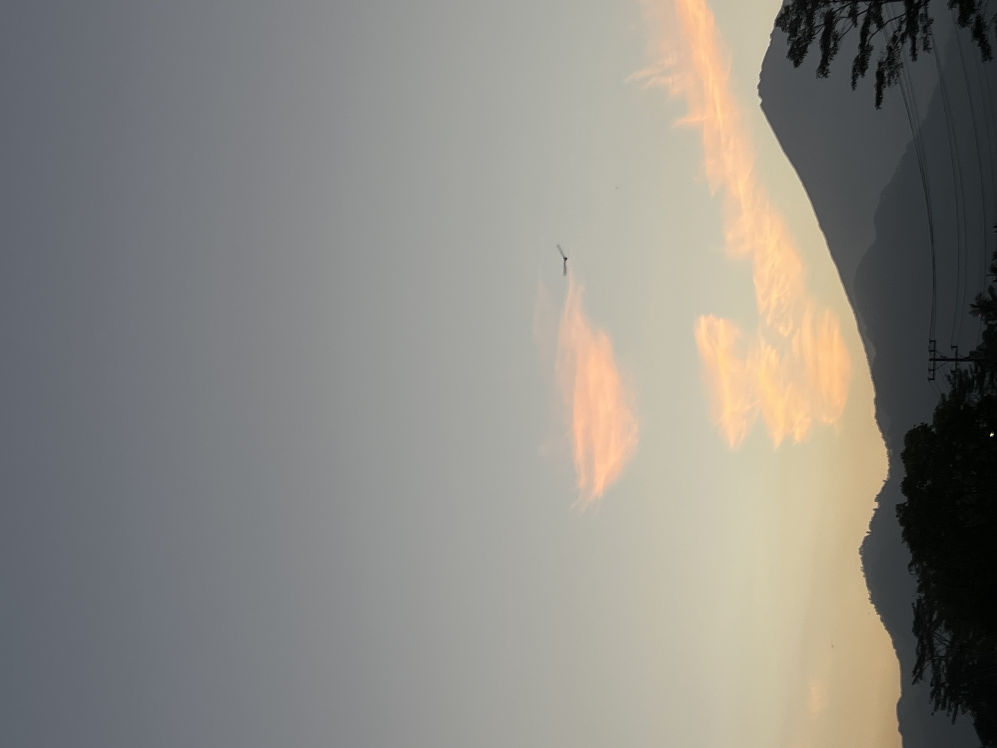 The image depicts a sunset or sunrise scene with a silhouette of mountains and trees in the foreground. The sky is a gradient of soft blues and oranges, with a few wispy clouds illuminated by the setting or rising sun. A single bird is visible in flight against the sky. The location is identified as Kalijoso, Indonesia. The overall atmosphere is serene and tranquil, capturing the beauty of nature during twilight hours. There are no people or specific man-made structures clearly visible, apart from what appears to be power lines in the lower part of the image.