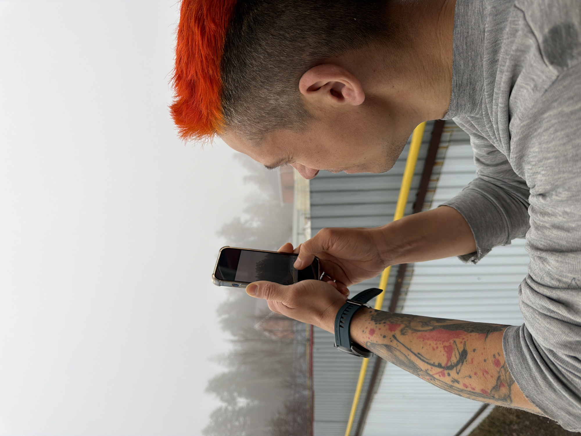 A person with an orange mohawk and a gray shirt is holding a smartphone and taking a photo of a hazy, overcast scene. The person has tattoos on their left arm and is wearing a smartwatch. The location appears to be outdoors, possibly overlooking a landscape obscured by fog. It is not possible to determine the specific location in Chaikovsky, Russia based solely on the provided image.