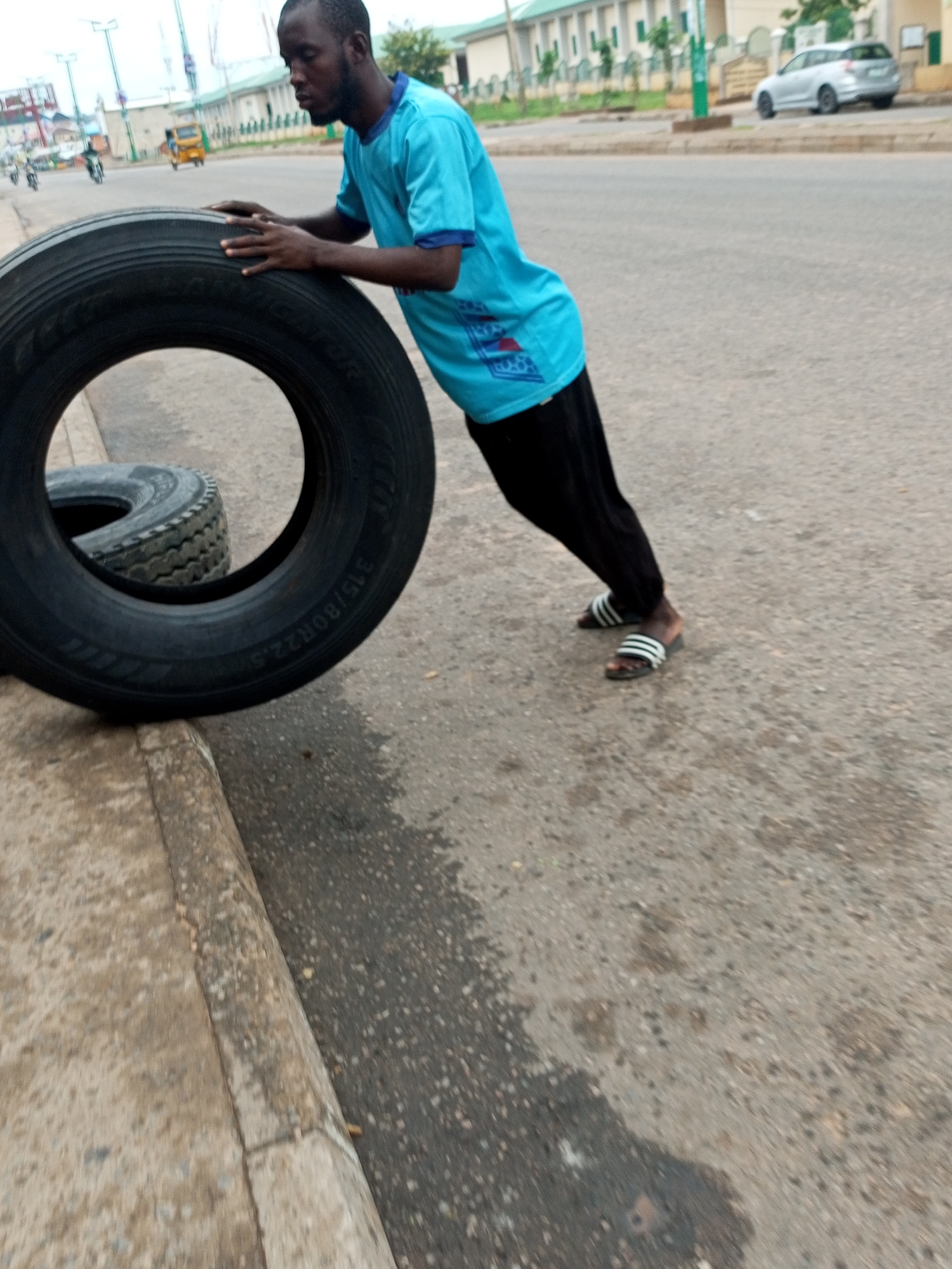 A young Black man wearing a blue t-shirt and black pants with white-striped sandals is performing a push-up against a large black tire. The tire is positioned on the edge of a sidewalk, with another tire partially visible underneath it. The man's body is angled upwards as he pushes off the tire. The background shows a street with vehicles, including a silver car, a yellow tricycle, and a few motorcycles. Buildings with green roofs and white walls line the street, suggesting an urban environment. The ground is paved and appears wet in places, indicating recent rain. The scene is set in Pantame, Nigeria.