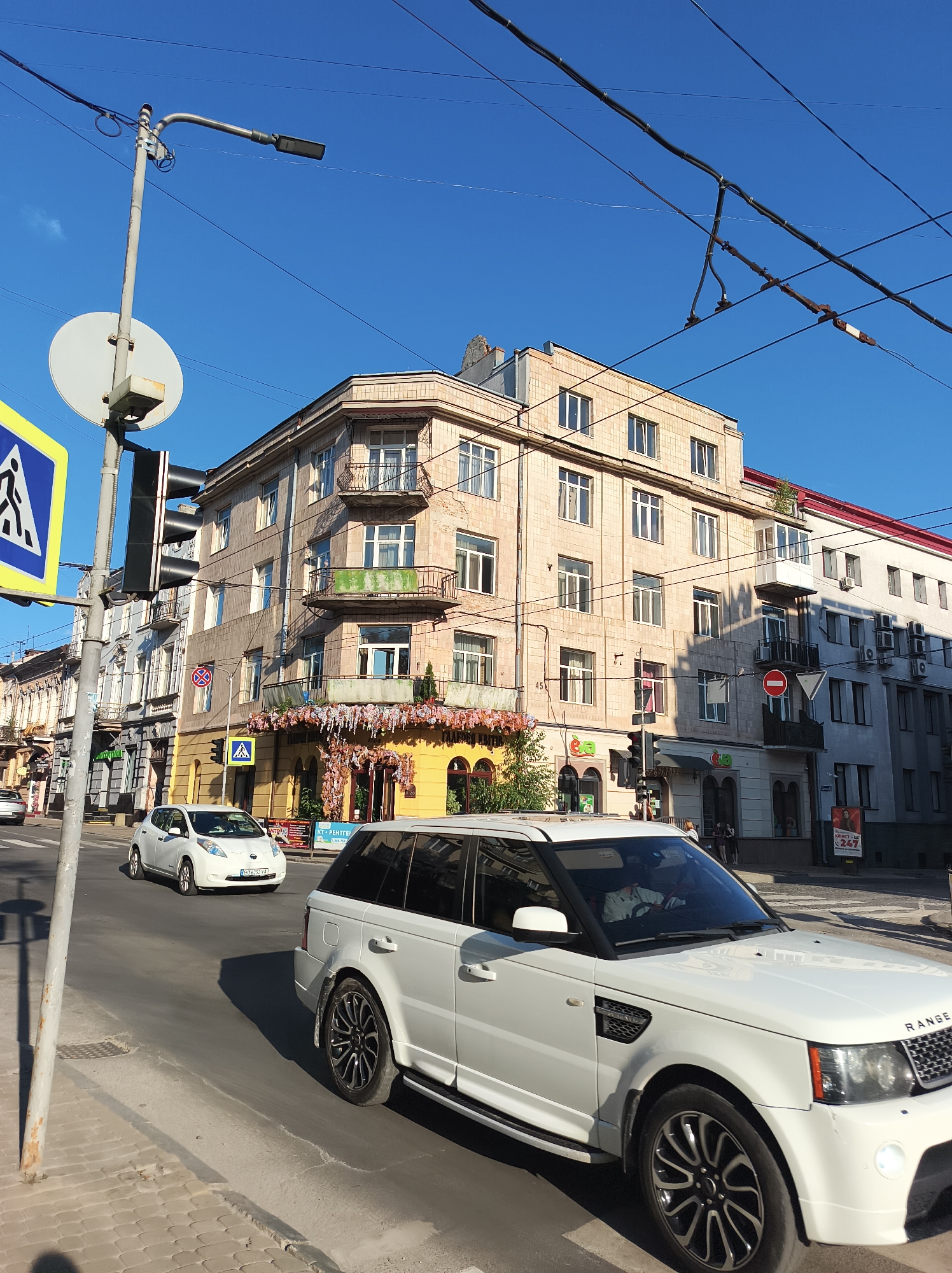 The image depicts a street scene in Ternopil, Ukraine, on a clear, sunny day. In the foreground, a white Range Rover is driving on the right side of the road. Behind it, a white Nissan Leaf is also in motion. The street is lined with buildings, with a prominent multi-story building with a light-colored facade and balconies taking center stage. This building features a yellow-colored facade at its base, decorated with floral arrangements. Across the street, other buildings are visible, including one with a red roof. Overhead, power lines and streetlights are present. Traffic lights and road signs, including a pedestrian crossing sign, are also visible. The scene appears to be a typical urban street with ongoing traffic.