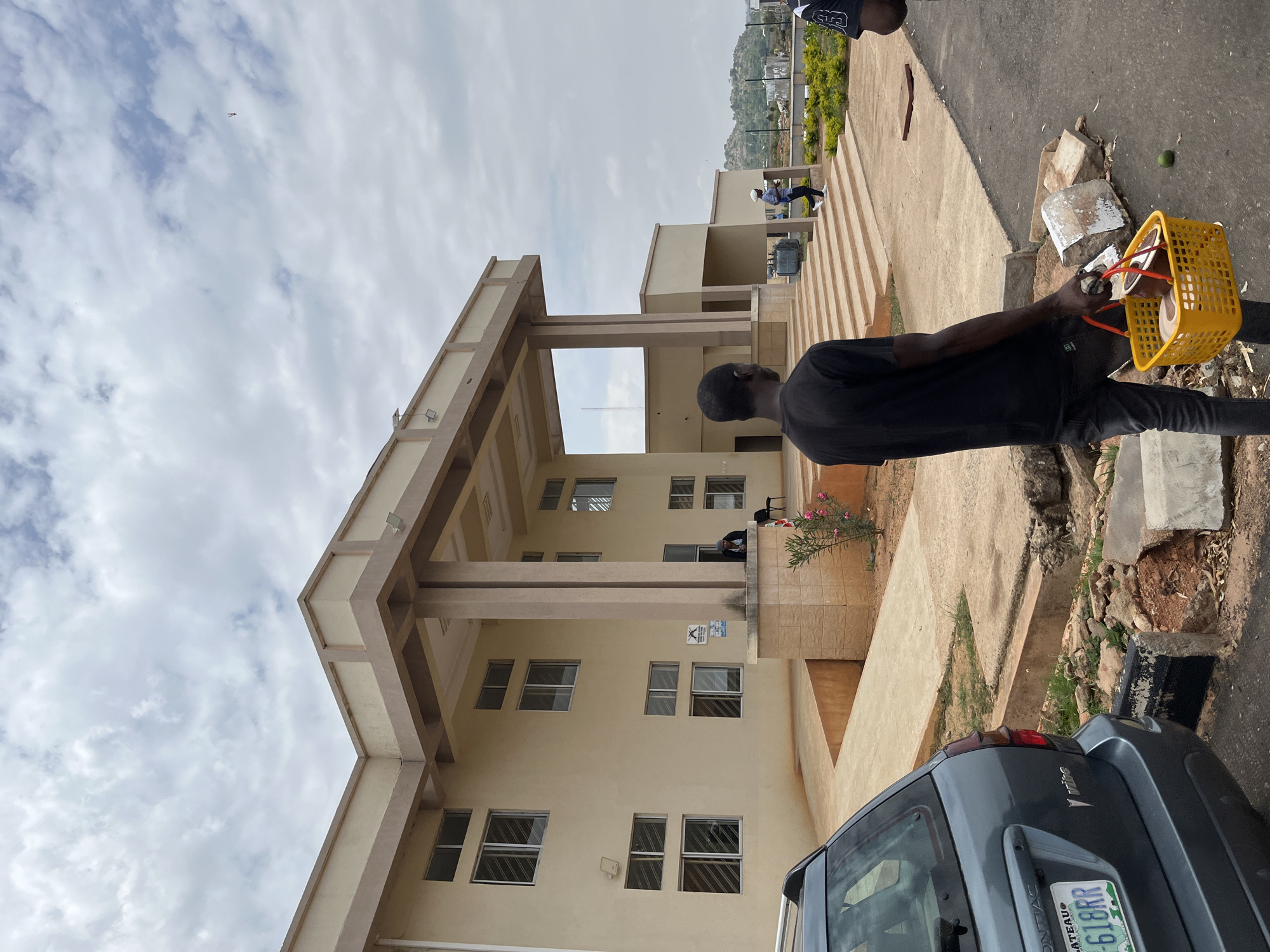 The image depicts a two-story building in Gwong, Nigeria. The building is beige with a prominent portico supported by columns. A man wearing a black shirt and black pants is standing on the right side of the frame, holding a yellow basket. A grey car is parked at the bottom right corner of the image. In the background, a hill can be seen. The sky is overcast with clouds. Several people can be seen in the background.
