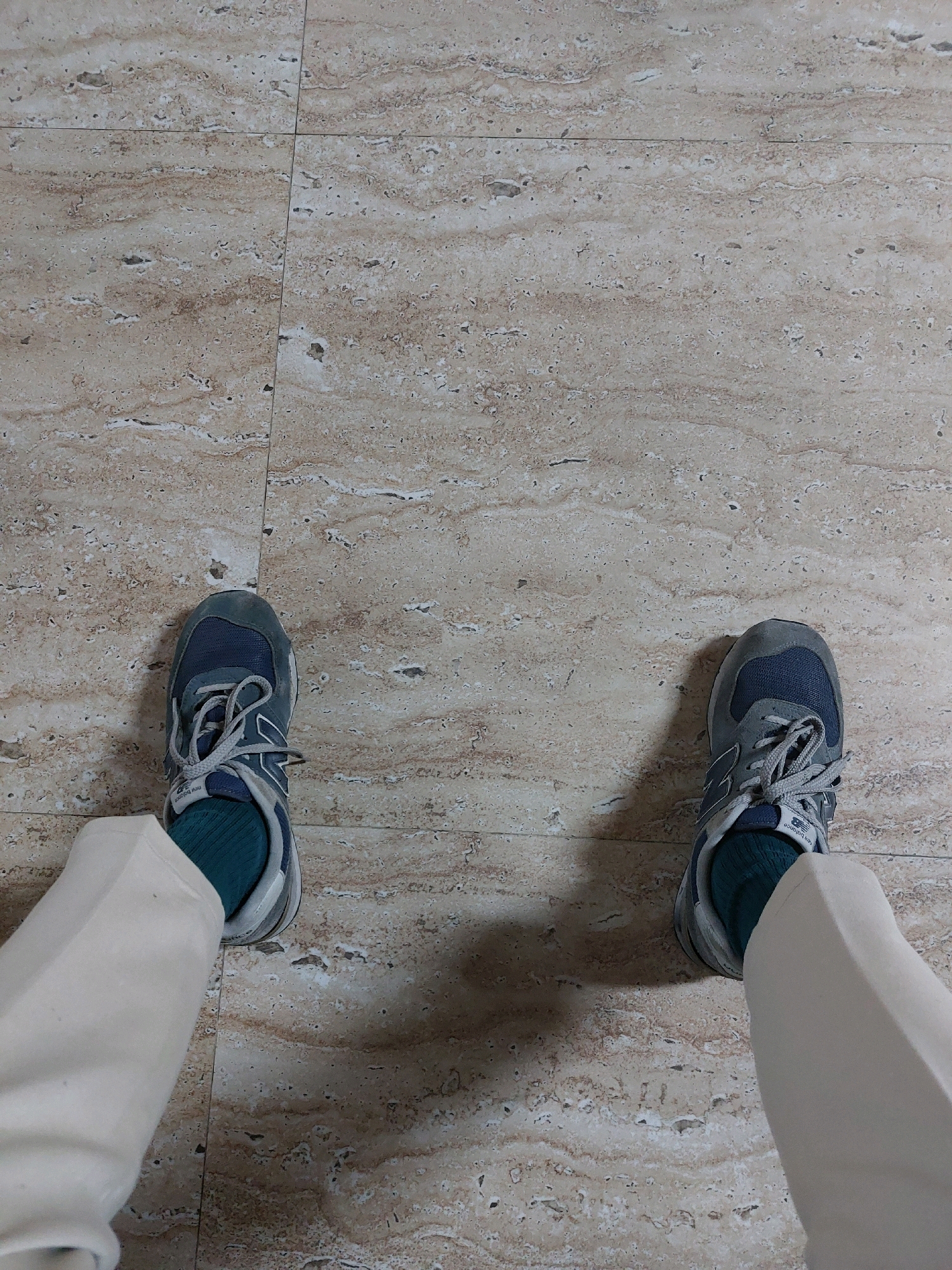 Two feet, clad in grey and blue New Balance sneakers and teal socks, are positioned on a beige travertine tile floor. The person is wearing light-colored trousers. The floor tiles are square with visible grout lines and a natural, veined pattern. The scene is indoors, likely a residential or commercial space in Wonokarto, Indonesia.