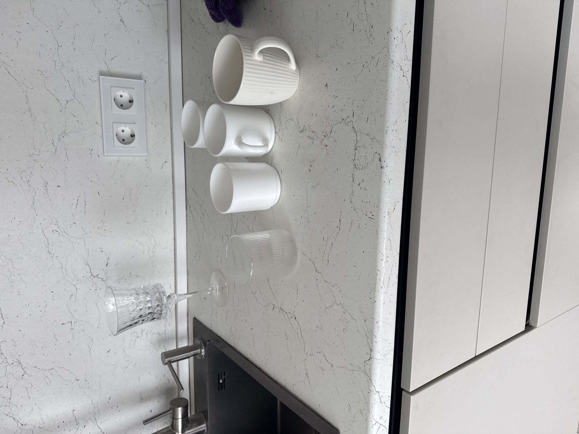 In a modern kitchen in Nizhnyarkeyevo, Russia, a white marble countertop displays several items. To the left, a stainless steel faucet extends over a dark sink. Next to the sink stands a clear, faceted wine glass. To its right, a ribbed clear glass is present. Further to the right, four white ceramic mugs are arranged in a cluster. Two are simple cylinders, one has a handle, and the fourth is a fluted mug with a handle. Behind the mugs, a purple cloth lies on the countertop. The wall above the countertop features a white double electrical outlet. The lower portion of the image shows light-colored kitchen cabinets.