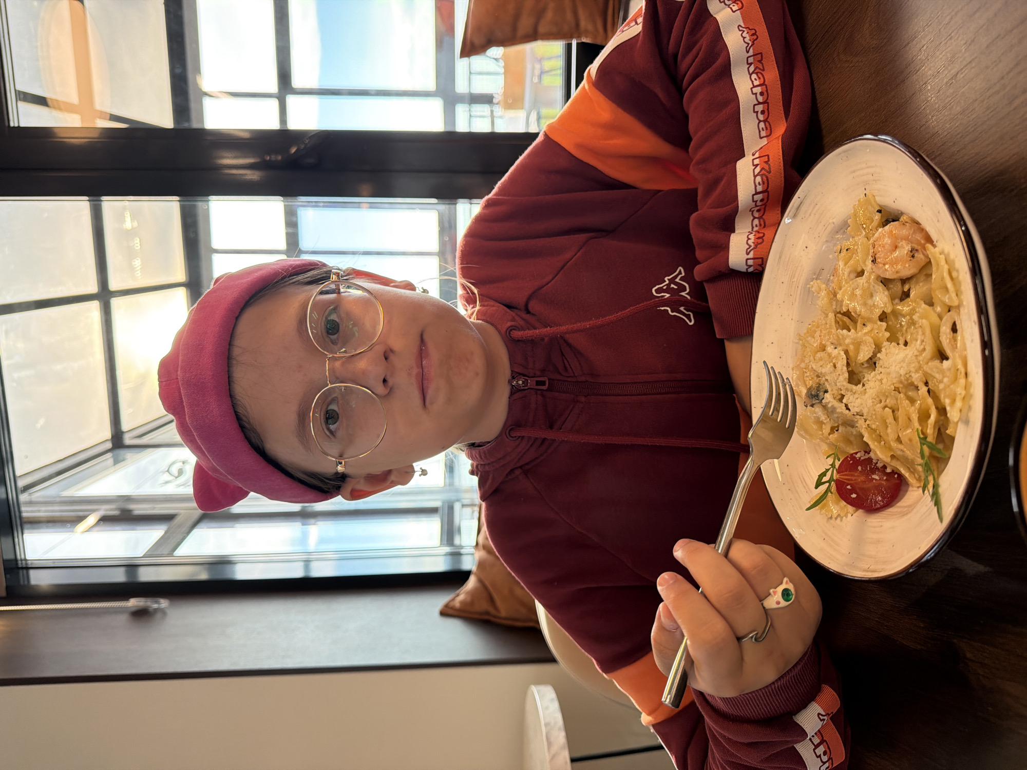 A young person is captured mid-meal at what appears to be a restaurant or cafe in Chaikovsky, Russia. They are positioned at a dark wooden table, looking directly at the camera with a neutral expression. The individual wears a pink beanie, round gold-rimmed glasses, and a maroon and orange hooded sweatshirt with the brand name "Kappa" visible in white text along a stripe on the sleeve, and the Kappa logo on the body.
In their right hand, they hold a silver fork, resting above a white plate filled with farfalle (bow-tie) pasta. The pasta is topped with a generous amount of grated cheese, a large shrimp, and a slice of red tomato garnished with a green herb. Two distinct rings adorn their right hand: one featuring a prominent green stone and another designed in the shape of a white cat with green eyes.
The setting is brightly lit by natural light streaming through a large window with multiple panes, visible behind the person. The window shows a clear, bright sky, suggesting it is daytime. A brown cushion or pillow is partially visible in the background, adding to the comfortable indoor ambiance. The overall impression is a casual dining experience.