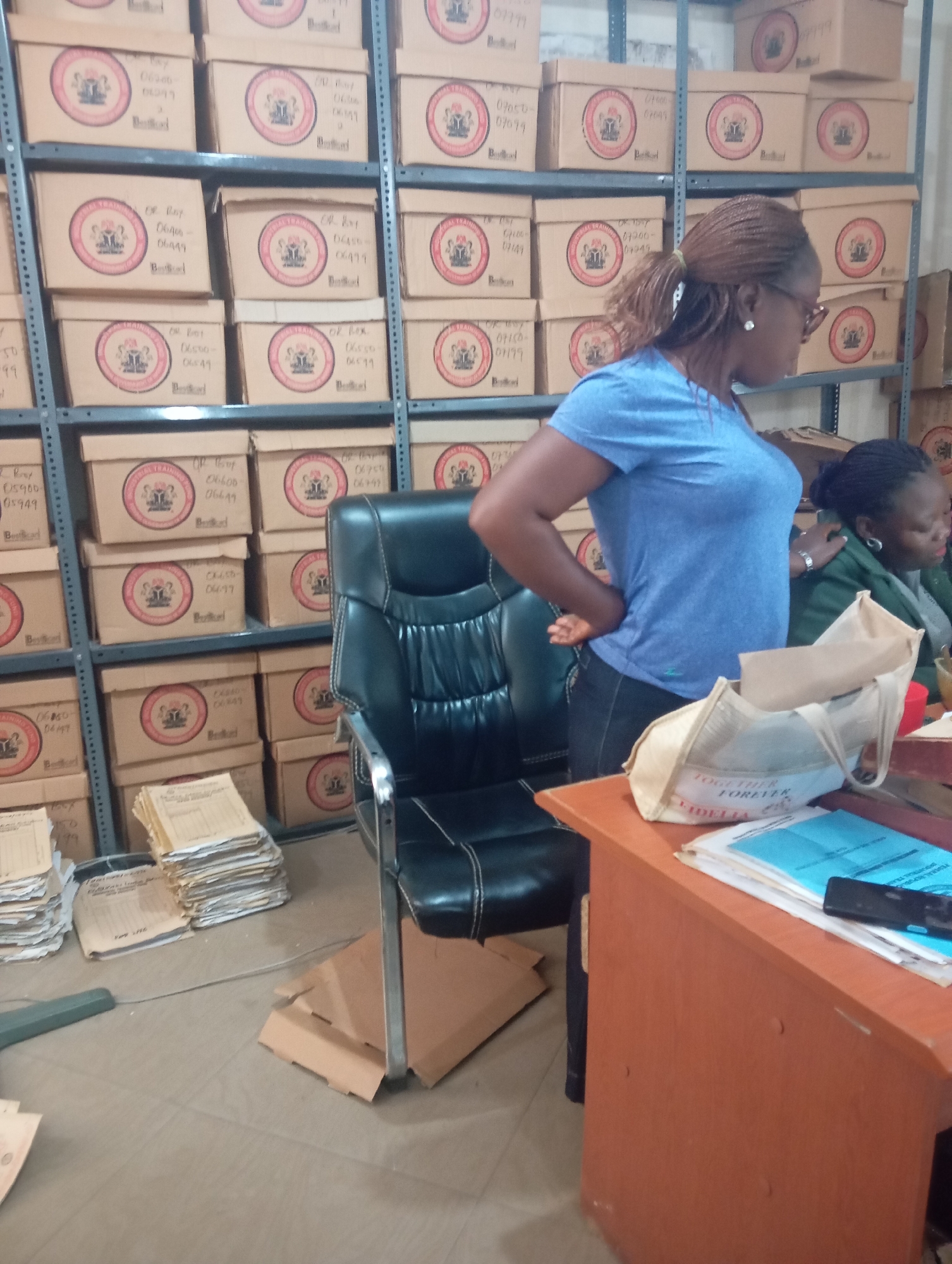 The image depicts an indoor scene, likely a storage room or office, filled with numerous stacked cardboard boxes. The boxes are uniform in size and bear a circular logo with text and an emblem. Some boxes have handwritten labels indicating numerical ranges and text such as "OR Roy" and "TRAINING". The overall impression is one of an organized but densely packed archive.
In the foreground, a woman with braided hair and glasses is partially visible, wearing a blue t-shirt and dark pants. She appears to be interacting with or observing a desk area. Another woman, wearing a green jacket and dark clothing, is seated behind the desk, looking down. On the desk, there are stacks of paper documents, a blue binder or folder, a mobile phone, and a beige tote bag with text that appears to read "TOGETHER FOREVER FIDELIA". A black executive chair is positioned between the women and the desk.
The environment suggests a functional space, possibly related to record-keeping or administration. The shelves holding the boxes suggest vertical storage, a common feature in archives or warehouses.
While the prompt mentions the city of Jos, Nigeria, there are no explicit visual cues within the image that definitively place the scene in Jos. The visual evidence is limited to the interior setting and the objects present.