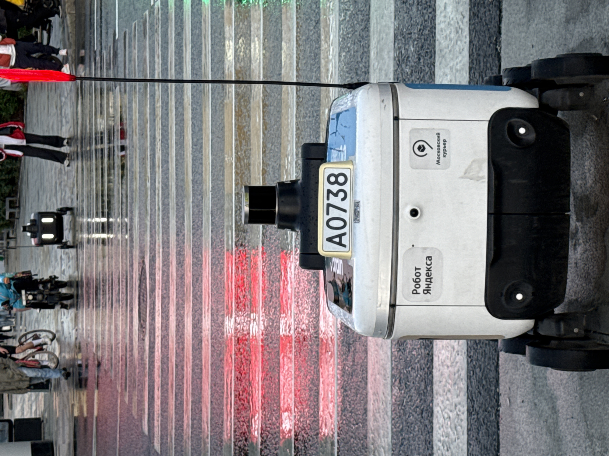 An autonomous delivery robot with the license plate A0738 is positioned at a pedestrian crossing in Moscow, Russia. The robot is white with a black base and has a lidar sensor on top. It displays logos for "Робот Яндекса" (Yandex Robot) and "Московский курьер" (Moscow Courier). The pedestrian crossing is wet, suggesting recent rain, with reflections of red and green traffic lights visible on the surface. In the background, several people are present, including individuals on bicycles and a person pushing a cart. Other autonomous robots are also visible in the background, moving across the street. The scene appears to be a busy urban environment with ongoing traffic and pedestrian activity.