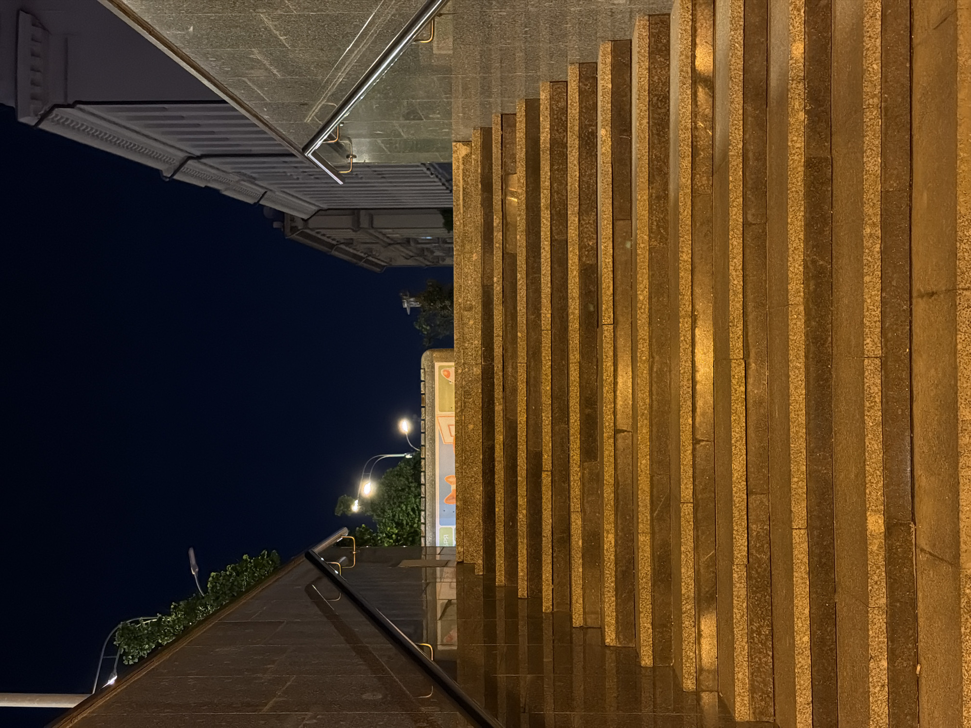 The image depicts a nighttime view of stairs in Moscow, Russia. The stairs are constructed from alternating bands of light and dark stone, illuminated by warm-toned lighting. A metal handrail runs parallel to the stairs, and a building is visible in the upper left corner of the frame. Streetlights are seen in the distance, amidst some greenery. The sky is dark.