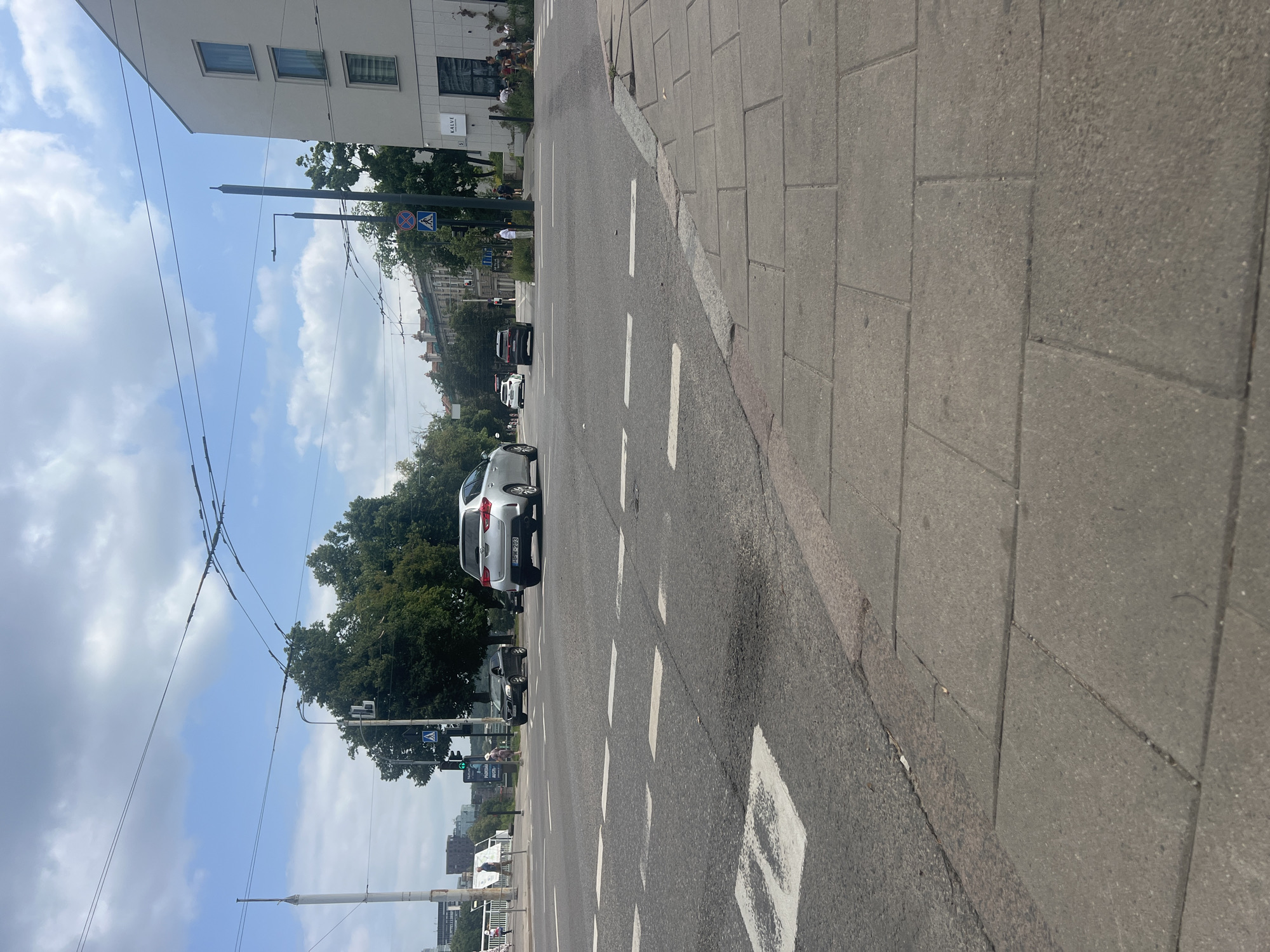 The image depicts an intersection in Vilnius, Lithuania. In the foreground, a paved sidewalk is visible, transitioning to a road with clear lane markings and a pedestrian crossing. Several vehicles are present at the intersection: a silver car with license plate HPM-203 is moving through, a dark SUV is stopped at the intersection, and other cars are visible in the background. A traffic light is illuminated green, indicating that vehicles can proceed. Trees line the street, and buildings can be seen in the distance, including a modern white building with windows. The sky is partly cloudy. There are a few pedestrians present, some near the buildings and others walking on the sidewalk. The scene appears to be a typical urban street view on a daytime.