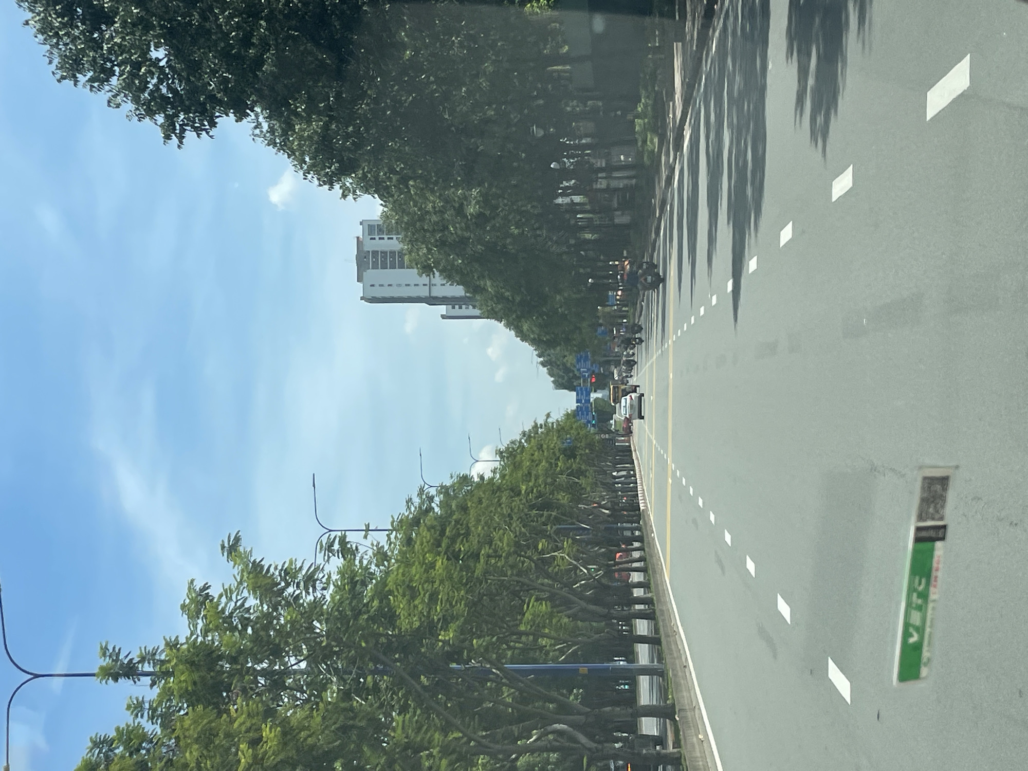 The image depicts a wide, multi-lane road in Thủ Đức City, Vietnam, lined with lush green trees on both sides. The road is paved and marked with white dashed lines indicating lanes. In the distance, there is a tall, modern building. The sky is blue with scattered white clouds, suggesting it is daytime. Several vehicles, including cars and motorcycles, are visible on the road. The trees form an avenue, creating a shaded canopy over parts of the road. There is a green and white sign on the road surface in the foreground.