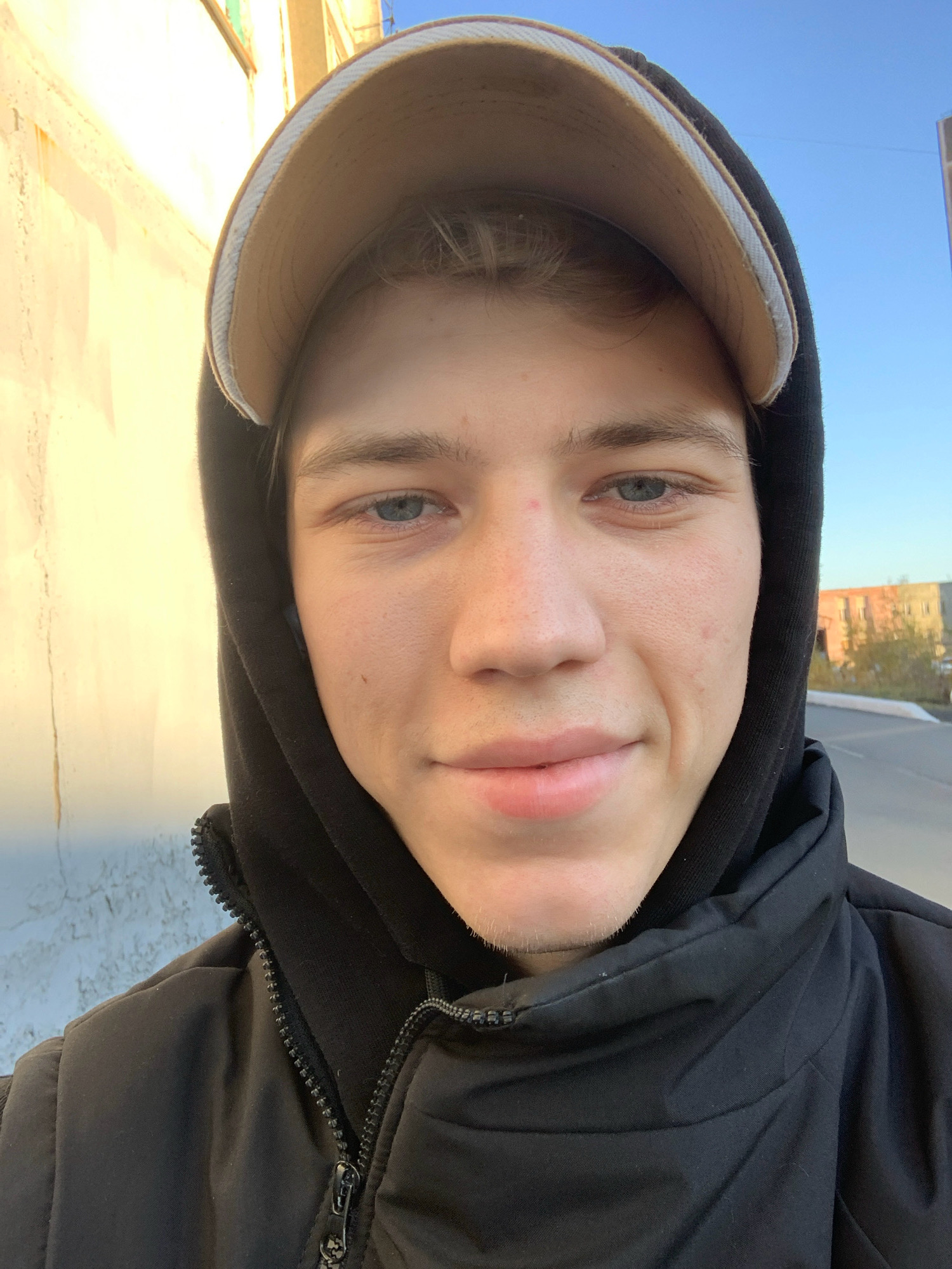 A young Caucasian male with light skin, blue eyes, and light brown hair is taking a selfie outdoors. He is wearing a black hooded jacket and a light brown baseball cap. The background shows a building on the left with yellow and white paint, and a clear blue sky. On the right, there is a street and some distant buildings. The overall lighting suggests it is daytime, possibly late afternoon given the warm tones on the building. The location is identified as Norilsk, Russia.