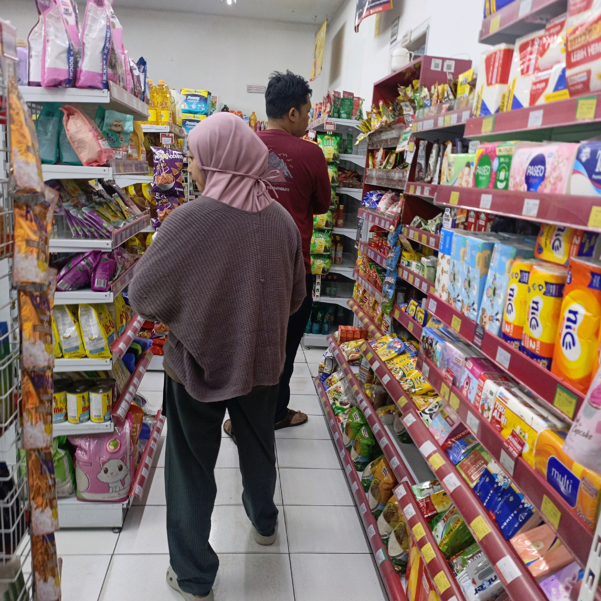 The image shows the interior of a convenience store in Salatiga, Indonesia, with two individuals browsing the aisles. The shelves are stocked with various consumer goods, including packaged food items, toiletries, and cleaning supplies.
In the foreground, a person wearing a pink hijab and a brown knitted sweater is facing away from the camera, looking at the products on a shelf to their left. They are wearing dark trousers and light-colored slip-on shoes. To their right and slightly behind them, another person wearing a maroon t-shirt with a graphic print is also facing away, looking at the shelves in front of them.
The store environment is well-lit, with white tiled floors and white walls. The shelves are made of metal and painted in red and white. The products displayed are diverse, featuring colorful packaging and prominent branding. The overall impression is of a typical retail setting within a neighborhood convenience store.