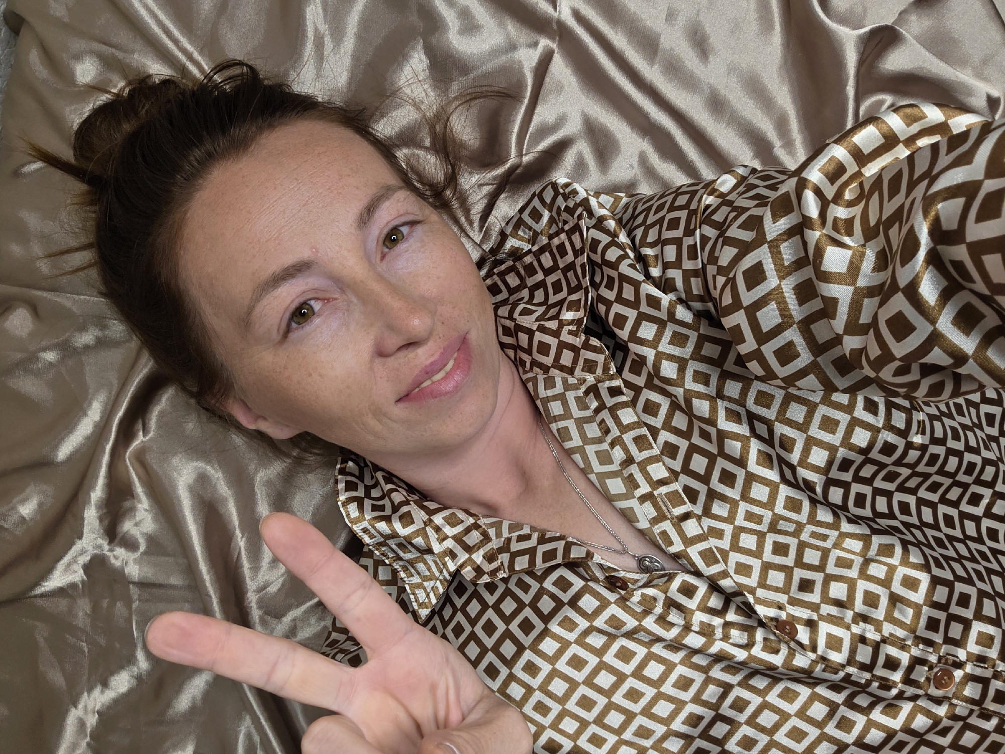 A woman lies on a satin bedsheet, her body angled towards the camera. Her hair is pulled up into a messy bun. She wears a gold and white patterned button-down shirt. Her right hand is raised, with her index and middle fingers extended in a peace sign. Her left hand is not visible. The lighting suggests an indoor setting, likely a bedroom. The overall color palette of the image is warm, with shades of brown and gold dominating.
