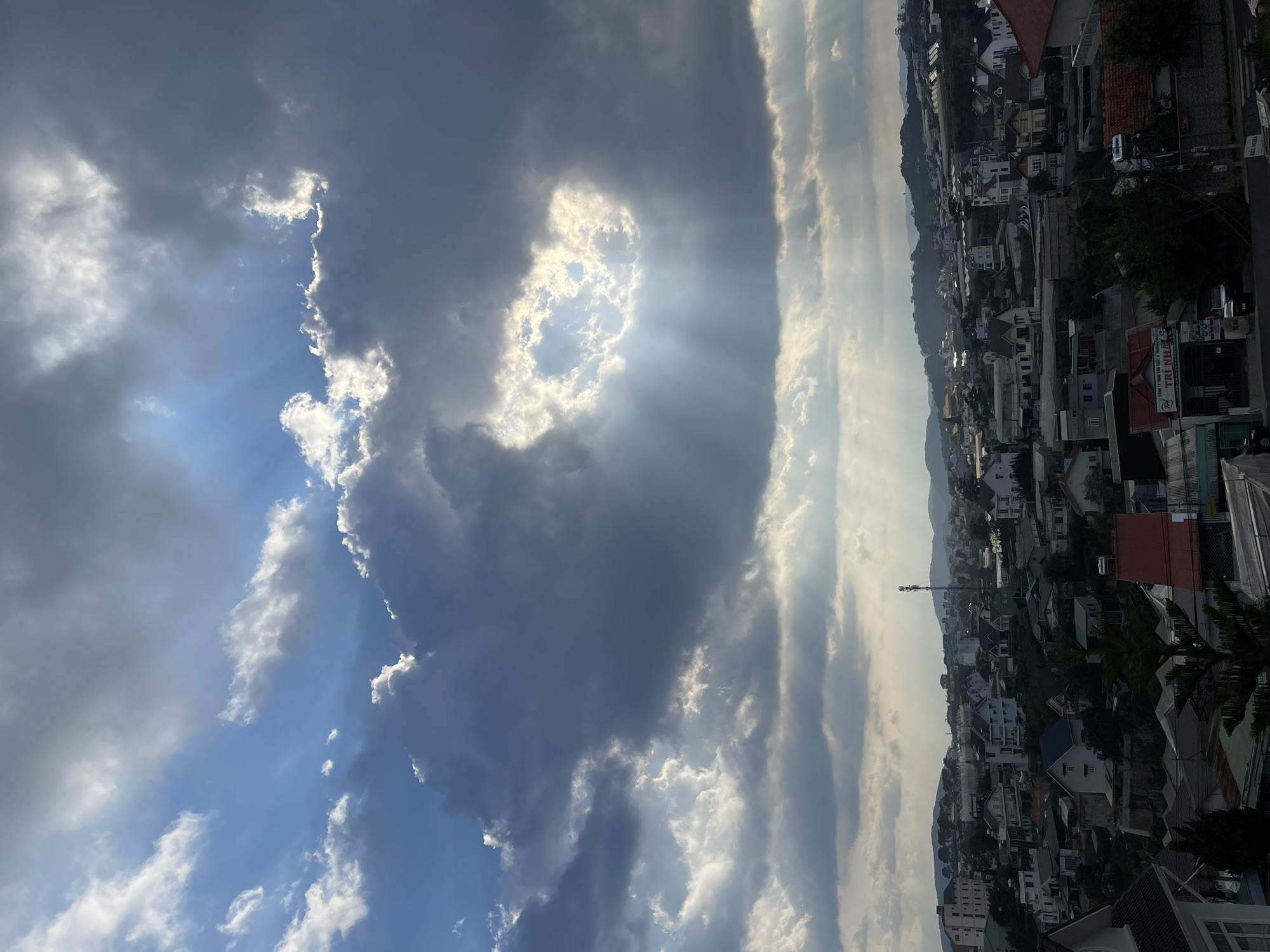The image shows a view of the city of Lâm Viên - Đà Lạt, Vietnam, under a cloudy sky with sun rays breaking through. The sky occupies the upper portion of the image, filled with a mix of dark and light clouds. Sunbeams radiate through a gap in the clouds. Below the sky is a landscape of houses, buildings, and vegetation stretching towards the horizon. The scene appears to be captured from an elevated position. The overall lighting suggests it is daytime.