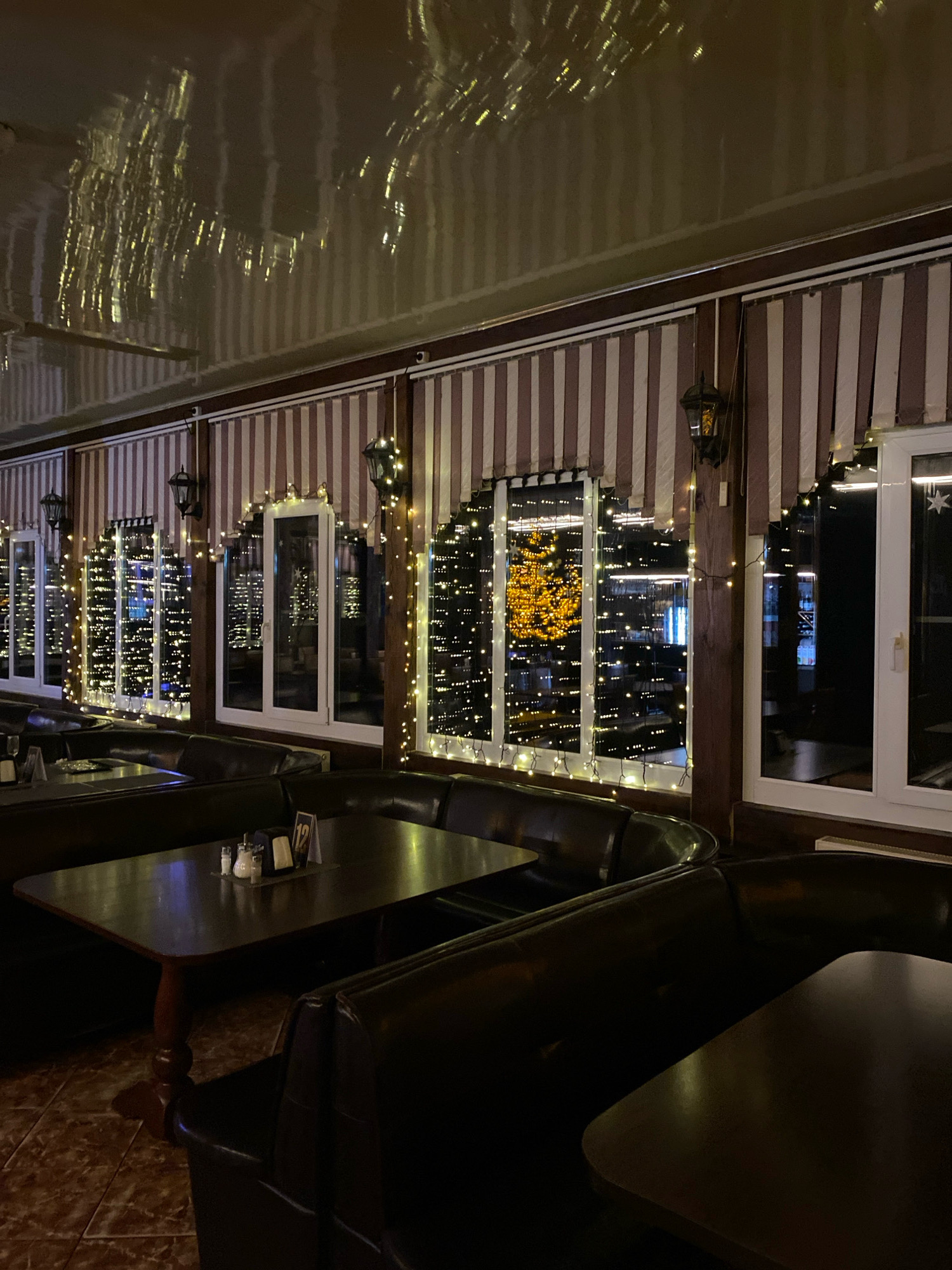The image depicts the interior of a dimly lit restaurant or cafe. The scene is dominated by dark leather booth seating arranged around wooden tables. The walls are lined with striped drapes, and windows offer a view of the exterior, illuminated by strings of warm white fairy lights. Some of the windows reflect the interior lights and decorations, creating a festive ambiance. Traditional lantern-style light fixtures hang on the walls. The floor is tiled with a mosaic pattern. The overall impression is one of a cozy and inviting dining space, likely decorated for a holiday or special occasion.