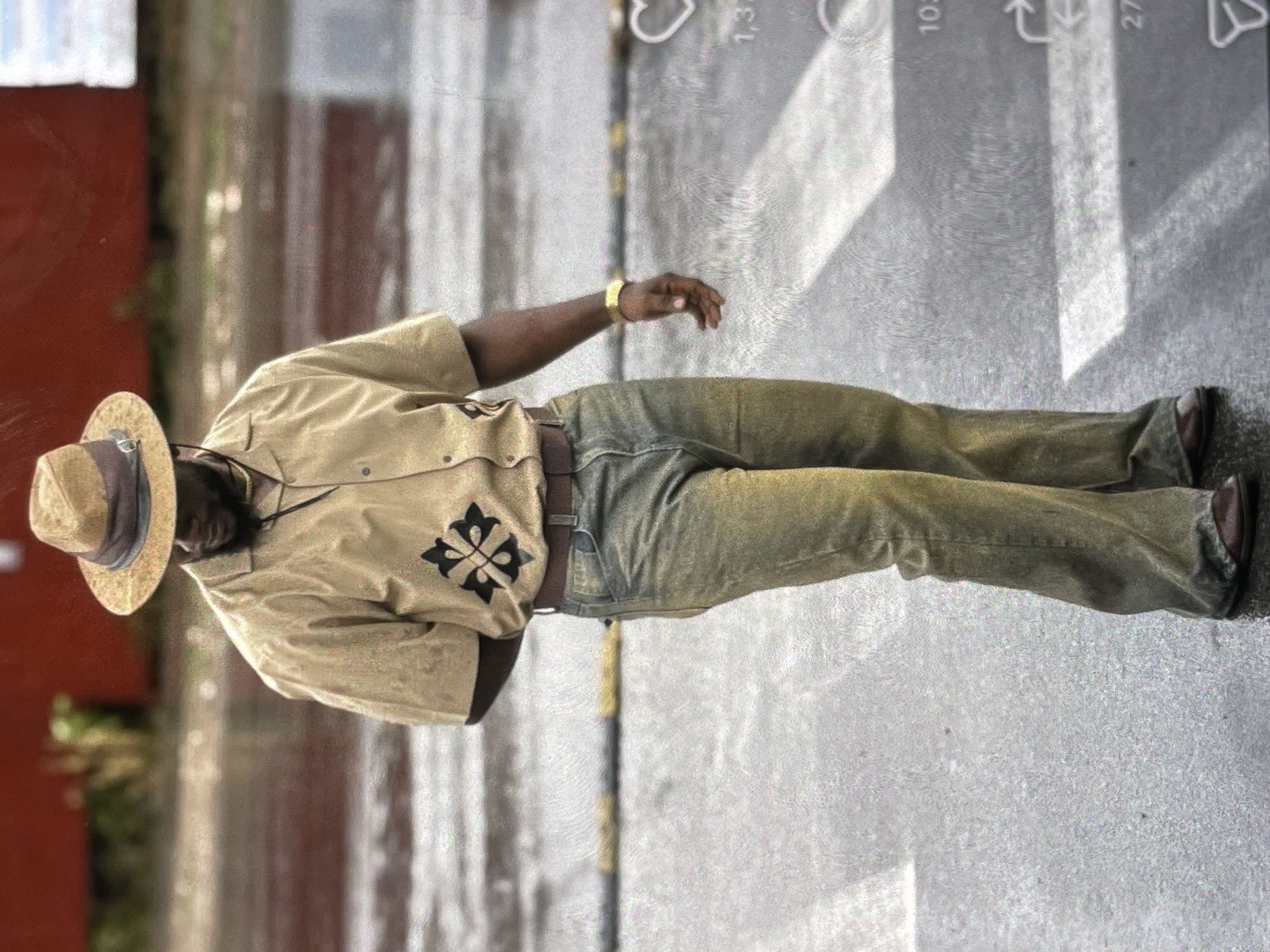 A full-shot photograph depicts a Black man from the chest down, standing on a wet asphalt surface with white and yellow striped crosswalk markings. The man wears a straw fedora hat with a dark band, a tan short-sleeved button-up shirt with a black embroidered emblem on the left side, a brown belt, faded greenish-brown denim jeans, and dark brown dress shoes. A gold-colored watch is visible on his right wrist. The background shows a blurred red structure. The lighting suggests an overcast day.