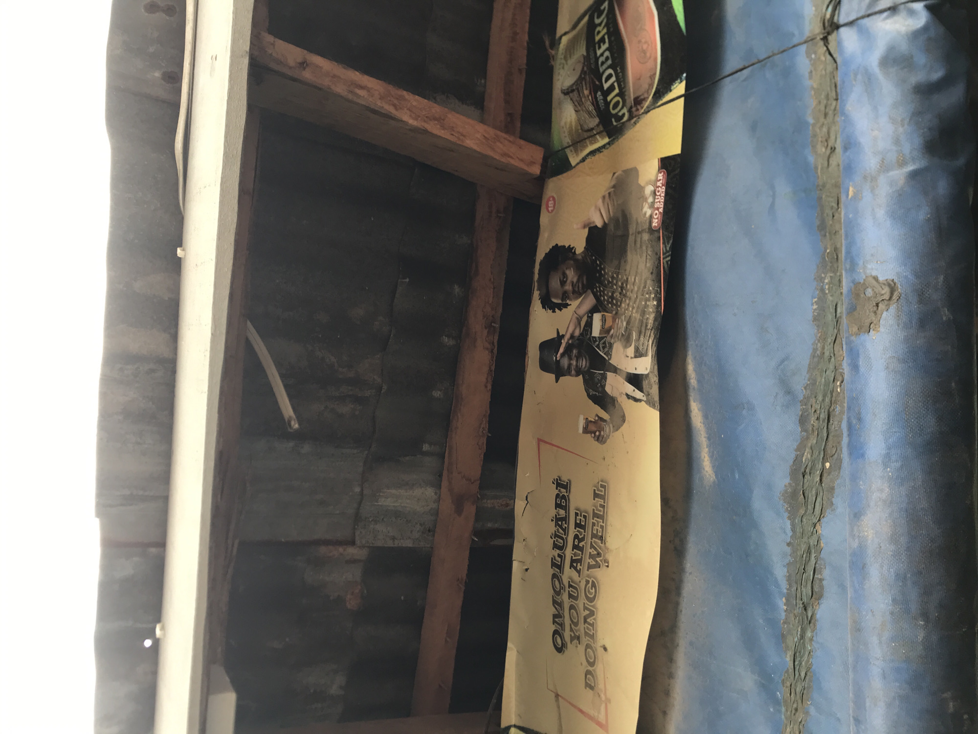 This low-angle image looks upwards into the ceiling structure of an interior space, likely a shelter or building in Jos, Nigeria. Exposed wooden beams and rafters form the framework, supporting corrugated metal sheets that appear to be roofing material. A bright white vertical beam or frame is visible on the far left, with a thin electrical wire running alongside it.
Prominently displayed on the right side of the frame is a yellowish-tan poster featuring two men. One man, wearing a hat, holds a glass and smiles at the camera, while another man behind him points. The poster contains the text "OMOLUABI YOU ARE DOING WELL", and partially visible details like "NO SUGAR ADDED" and "18+" suggest it's an advertisement for a beverage. Above this poster, part of a "GOLDBERG" brand label is visible, indicating another advertisement.
To the right of the posters, a blue tarp-like material is stretched across the ceiling, featuring a dark, rough, and uneven seam running across its surface. The lighting is uneven, with an intensely bright, overexposed area on the far left, indicating a strong light source, possibly an open side or window. The overall setting suggests a rustic or utilitarian construction, utilizing readily available materials, with commercial advertisements integrated into the environment.