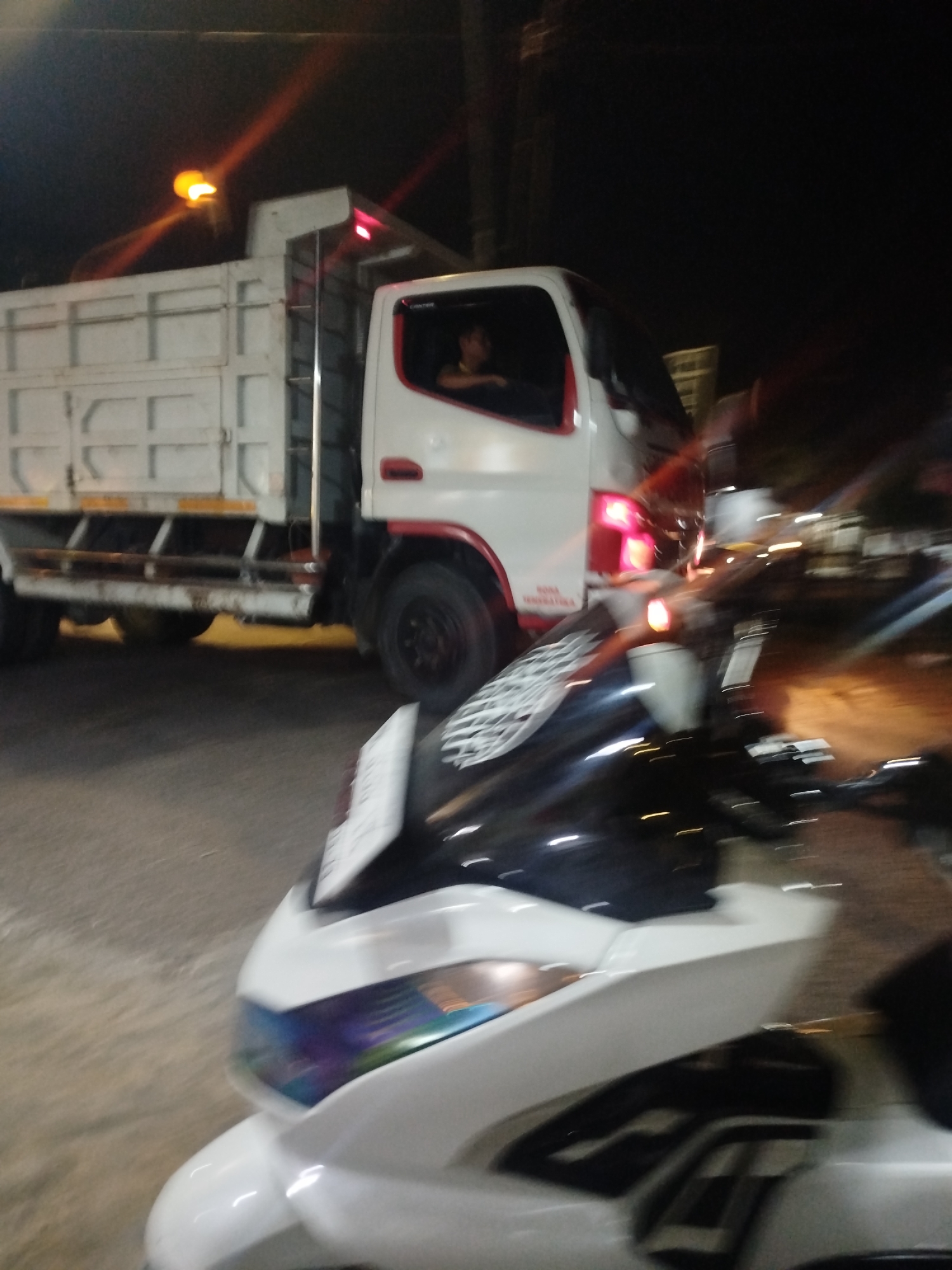 A white truck with a white cargo box is parked on a street at night, with its headlights on. A person is visible through the driver's side window. In the foreground, the front of a white motorcycle is visible, with its headlights on and reflecting on the road. The scene appears to be in Tonjung, Indonesia, based on the context.