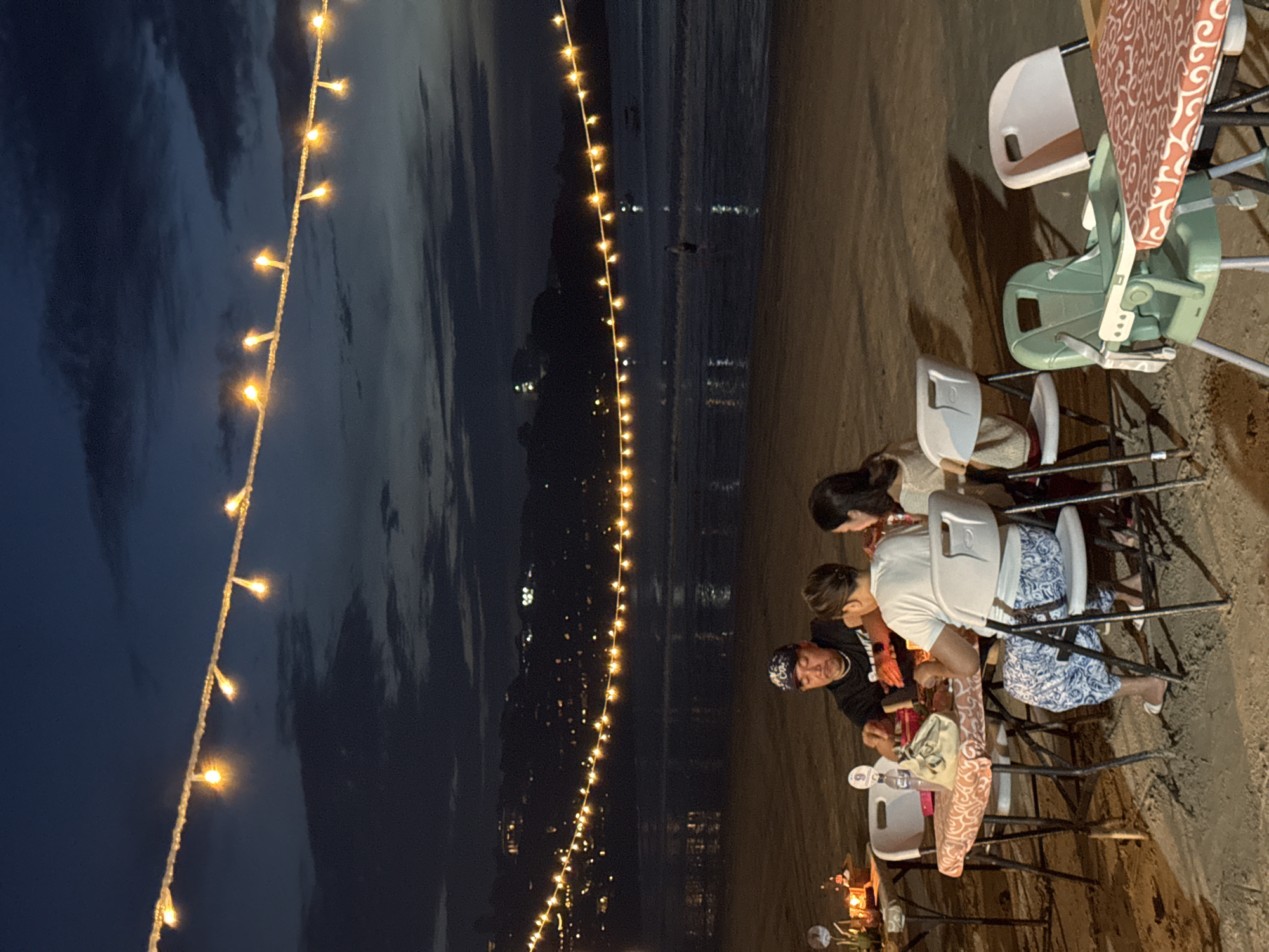 The image, taken at dusk or night, shows a group of people gathered on a sandy beach in Kuta Selatan, Indonesia, illuminated by string lights. The photograph is oriented vertically, with the beach and people on the right side and the dark sky and ocean on the left.
In the foreground, several individuals are seated around tables and chairs set directly on the sand. A man wearing a black long-sleeved shirt and a bandana is visible on the left. Next to him, another person is seated in a white t-shirt and blue and white patterned shorts or pants. A woman with dark hair is also part of the group. White plastic chairs are used, some featuring patterned red and white seat cushions. A green baby high chair or booster seat is also visible, suggesting a family setting.
Overhead, a long string of warm yellow lights stretches across the scene, providing ambient illumination and creating a festive atmosphere. The background reveals the dark ocean, with faint reflections on its surface and distant city lights twinkling along the coastline. The sky is dark, indicating evening hours. The overall scene depicts a relaxed outdoor dining or social gathering by the sea. No legible text is visible in the image.