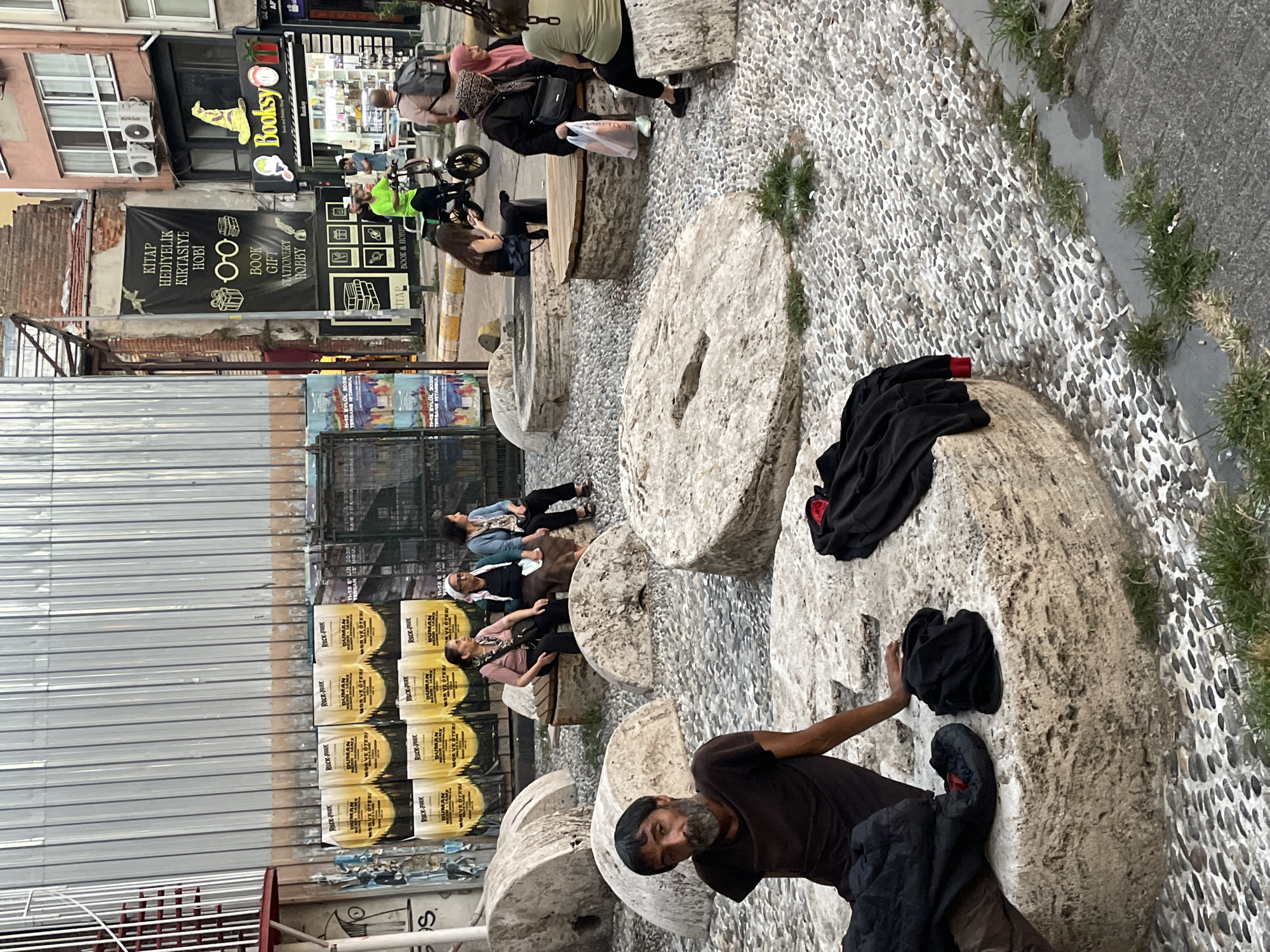 In Kadıköy, Turkey, a group of people are gathered in an outdoor area paved with pebbles and scattered with large, circular stone millstones. In the foreground, a man with a beard sits on one of the millstones, looking directly at the camera. Clothing, including a black jacket and pants, are placed on or near the millstones. Several other individuals are seated on or around the millstones, some engaged in conversation. A woman in a pink scarf is seated on a millstone, while another woman in a white headscarf and blue cardigan sits next to her on a wooden bench. In the background, a man wearing a bright green shirt rides a bicycle. Various posters and advertisements are displayed on a building wall. A shop sign with the word "Booksy" is visible, indicating a commercial establishment. The overall scene depicts a public space with people relaxing and interacting amidst the unique stone structures.