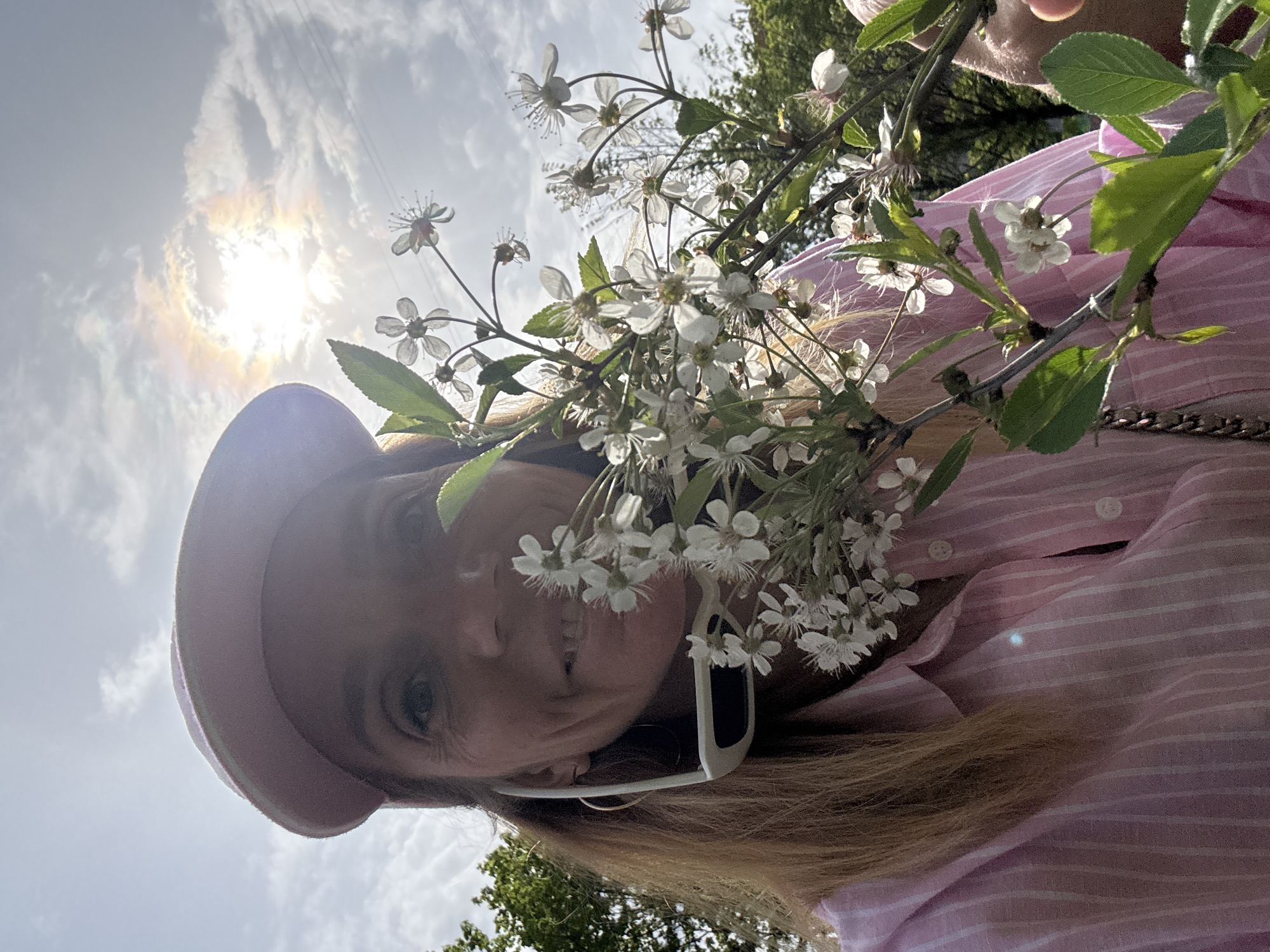 The image shows a woman in a pink striped shirt and a pink hat, holding a branch of white flowering tree blossoms. She wears white-framed sunglasses. The background includes a sky with the sun and clouds, and trees. The location is not possible to determine from the image, and there is no evidence of Saint Petersburg, Russia.