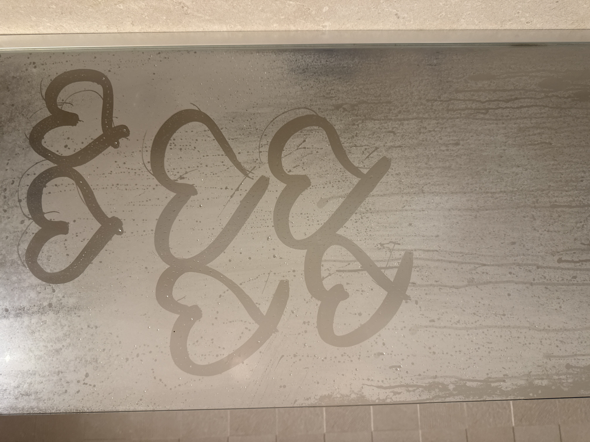 A steamy glass panel, likely a shower door, occupies the majority of the frame. Numerous heart shapes have been drawn or wiped into the condensation on the glass. There are at least seven distinct heart outlines visible, predominantly on the left and central portions of the panel. These hearts appear hand-drawn, clearing away the fog to reveal the clearer glass beneath, which appears darker than the surrounding misty areas.
The entire glass surface is covered in a dense layer of condensation, characterized by countless tiny water droplets and some visible streaks where water has begun to run down. The right side of the panel shows more pronounced vertical drip trails, indicating the natural flow of condensation.
Above the glass panel, a light-colored, possibly beige or off-white, tiled wall is visible, framed by a thin silver-colored border where it meets the glass. Below the panel, a similar tiled surface, likely the shower floor or a lower wall section, is visible, extending across the bottom of the image.
The scene suggests a bathroom setting, specifically the interior of a shower enclosure after a hot shower has generated significant steam and condensation. The act of drawing hearts on the fogged glass conveys a playful or affectionate sentiment. No discernible text is present, only the heart shapes. The lighting is soft and even, typical of indoor artificial light.