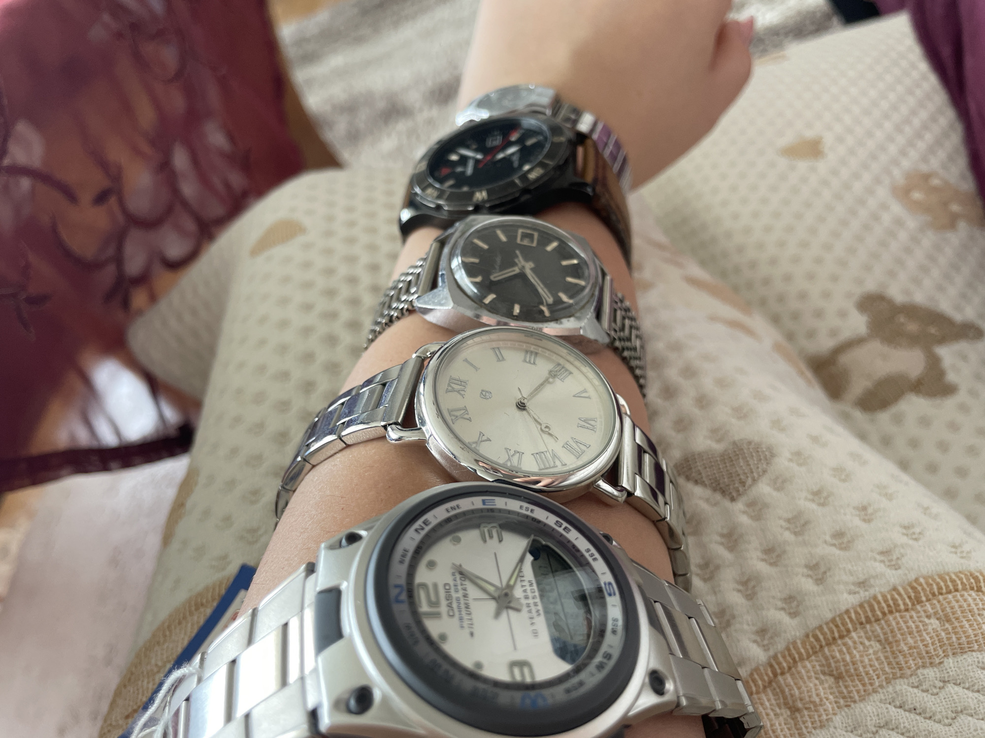 Four wristwatches are visible on a person's arm, resting on a patterned fabric. The watches are positioned from left to right. The first watch, a silver Casio, has a digital display and analog hands. The second is a silver analog watch with Roman numerals. The third, a black analog watch, is partially obscured. The fourth watch, also black, is the most distant and its details are less clear. The fabric underneath is light-colored with a repeating pattern of small, brown shapes. The background is blurred, but suggests an indoor setting with a hint of a wooden surface and floral upholstery. The image is taken from a slightly elevated perspective looking down at the arm and watches. There is no discernible information about the location in Zlatograd, Bulgaria.