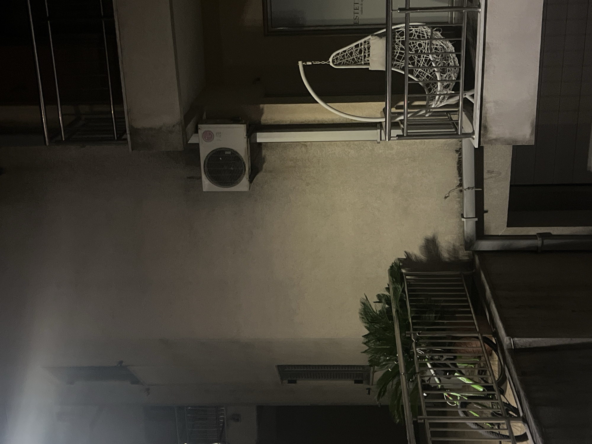 View of an outdoor urban scene at night, likely in Budva, Montenegro. An air conditioning unit is mounted on the wall. A white, hanging, egg-shaped chair is seen on a balcony. Below, a stairway is lined with plants. The location appears to be residential.