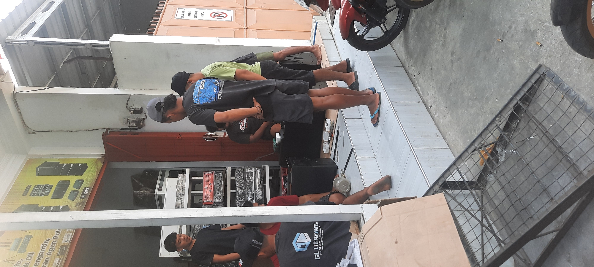 In Selopuro, Indonesia, a group of men stand outside a shop. One man in a black t-shirt with "GL LIGHTING PROFESIONAL LIGHT" printed on it is partially visible. Another man wears a grey baseball cap and a black t-shirt with a graphic design. A third man in a lime green t-shirt and dark shorts stands to his right, and a fourth man with a black hat and t-shirt is seated. They appear to be gathered around what looks like audio equipment, with several amplifiers and speaker boxes stacked on shelves inside the shop and on the ground outside. A red motorcycle is parked to the right of the frame. The shop has a light-colored exterior with an open doorway. A sign with Indonesian text and a circular "No Parking" symbol is visible on the right side of the building. A metal grate structure is in the foreground, obscuring part of the ground. The scene suggests a commercial setting, possibly a sound system store or repair shop.