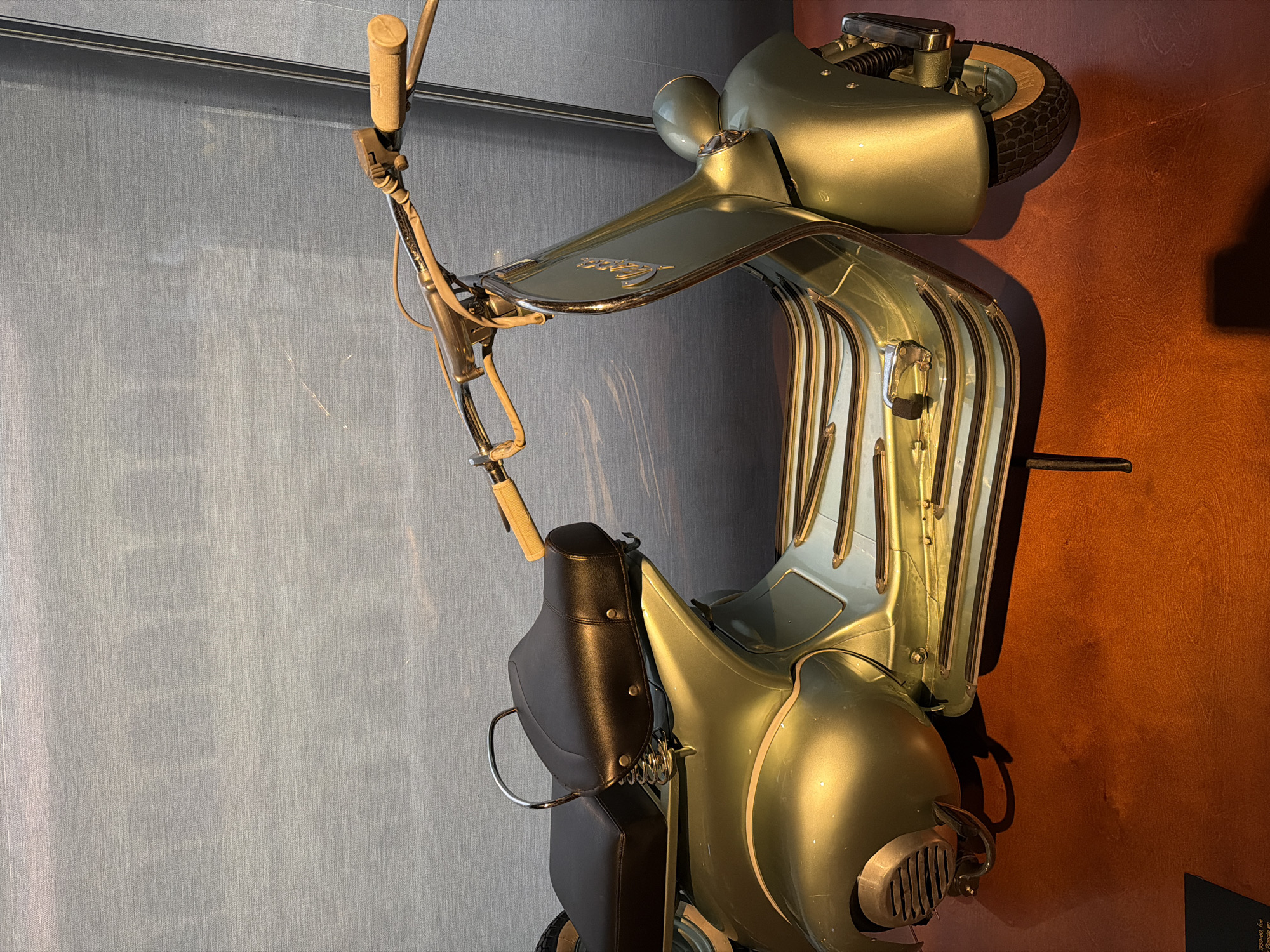 The image shows a vintage light green Vespa scooter mounted on a wall, likely for display purposes. The scooter features black leather seats and accessories, and its chrome accents are visible. The handlebars have wooden-looking grips. The background is divided into two sections: one is a light blue textured wall, and the other is a dark reddish-brown wall. This image was taken in San Sebastian, Spain.