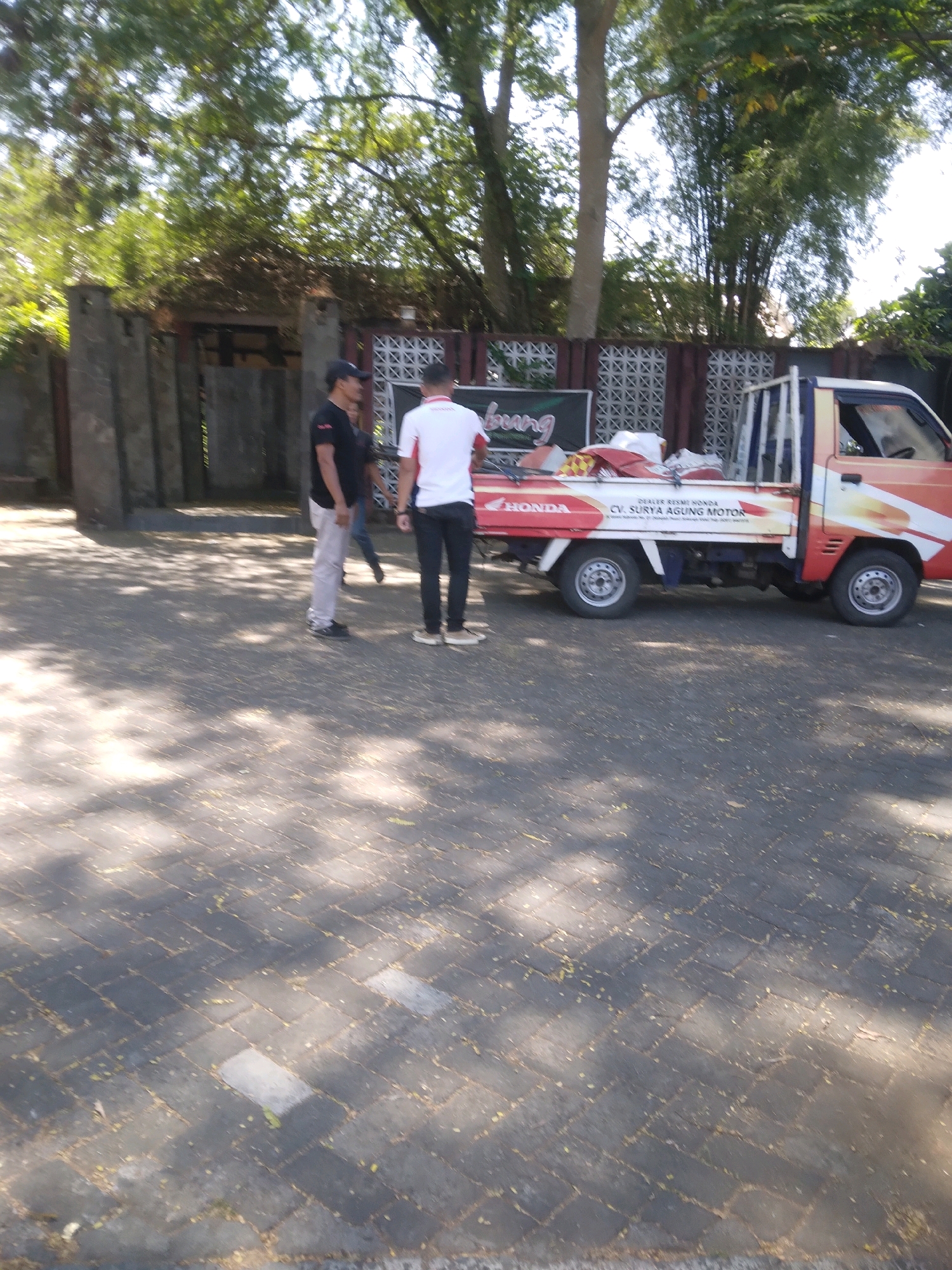 In Grendeng, Indonesia, a truck is parked on a paved lot near a building. Two men are standing next to the truck, one in a black shirt and the other in a white shirt with red accents. The truck is orange and white and has "HONDA" and "CV. SURYA AGUNG MOTOR" written on its side. The area is outdoors and sunny, with trees and shadows visible.