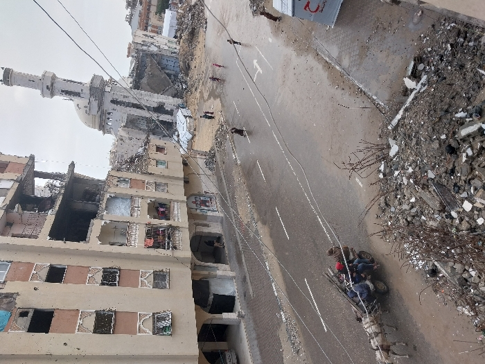 This image depicts a street scene in Khan Yunis, Palestinian Territory, characterized by extensive urban destruction under an overcast sky. In the foreground, a substantial pile of concrete rubble and debris marks the immediate edge of the frame. A wet, asphalt road with faint white dashed lane markings extends through the mid-ground. On the left side of the road, a light-colored horse pulls a two-wheeled cart carrying four individuals and several long, slender items, moving leftward. Further along the street, several pedestrians, including three children, are visible walking on the wet asphalt. On the right side of the street, a white structure bearing a red crescent emblem is partially visible.
In the background, a multi-story building with a light-colored facade stands on the left, displaying significant structural damage to its upper floors, including exposed interiors, missing sections, and shattered windows; laundry is visible on one of its balconies. Dominating the center-right background is a large mosque with a light-colored dome and a tall minaret, both extensively damaged. Large portions of the mosque's walls and superstructure have collapsed, leaving a significant debris field around its base. Additional collapsed structures and debris are visible further in the background, indicative of widespread devastation.