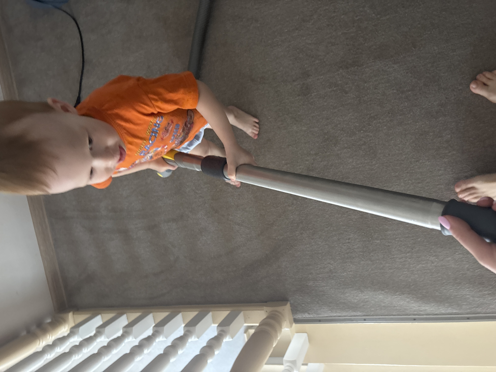 A young child wearing an orange shirt holds the metal wand of a vacuum cleaner, as an adult, visible only from the hand and feet, assists. The setting is indoors on a carpeted floor near a white wooden railing. The specific location in the city of Ферма, Russia, cannot be determined from the image.