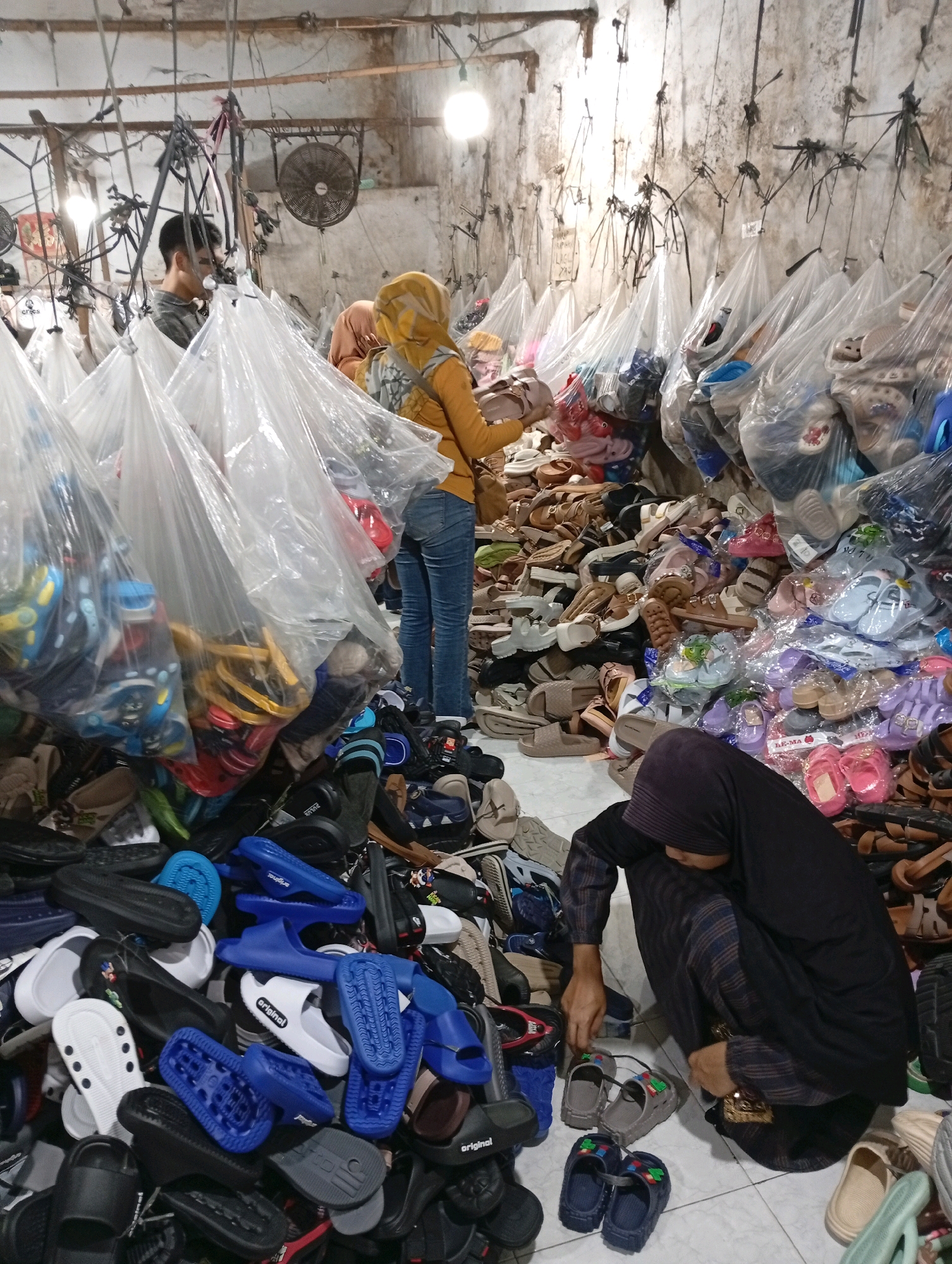 In a cluttered storage room in Babakan, Indonesia, a man stands partially visible behind plastic bags filled with shoes, which hang from the ceiling. To his right, a woman in a yellow hijab and orange jacket sorts through a pile of footwear. In the foreground, a woman in a dark hijab crouches and examines a pair of sandals. The room is filled with numerous pairs of shoes, many of which are in clear plastic bags, and others are piled on the floor. The walls are rough and dirty, with exposed pipes and wiring. A fan is mounted on the back wall. The overall impression is one of a busy, disorganized retail or storage space for footwear.