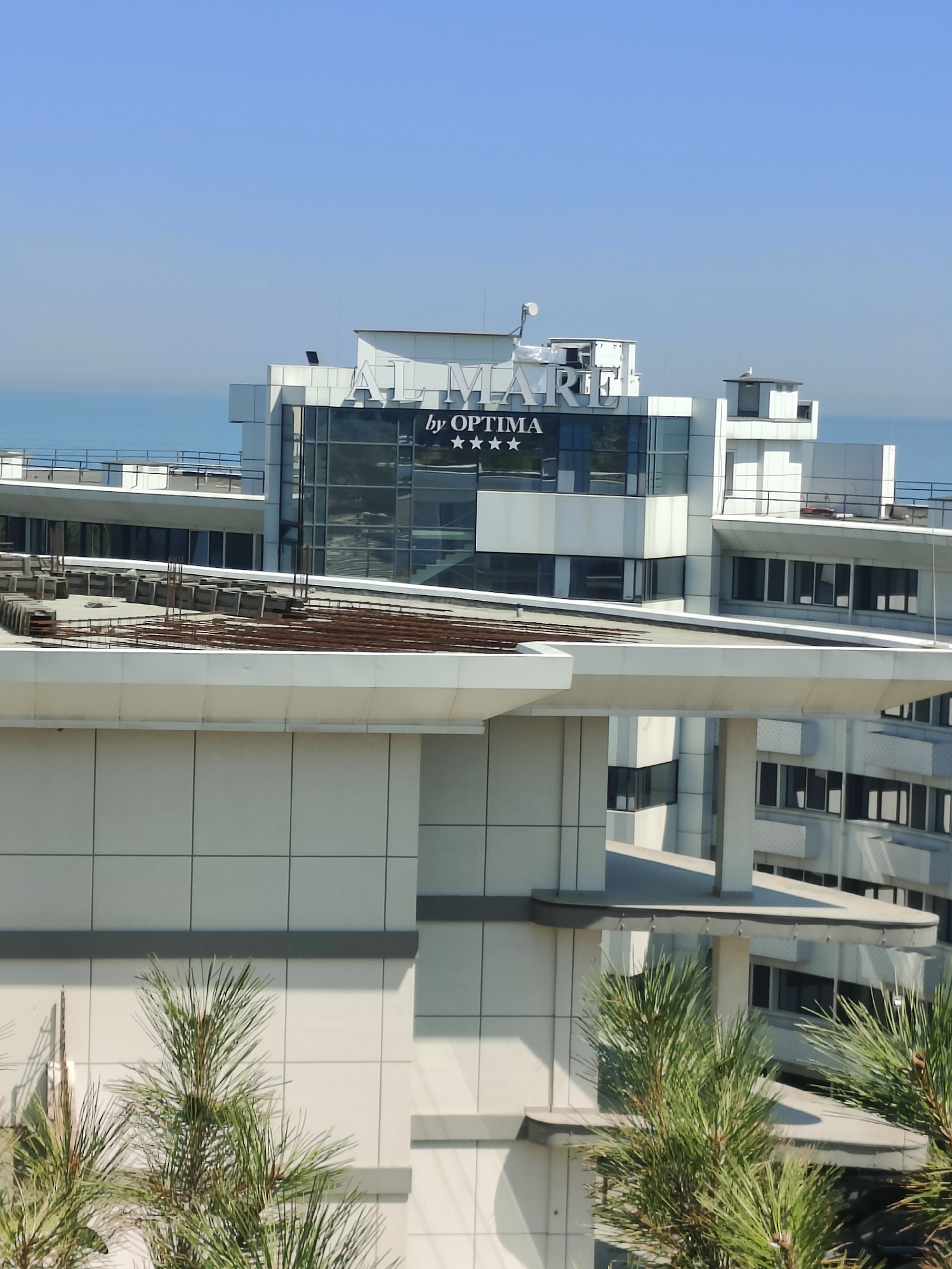 The image shows a modern hotel building on a clear, sunny day, with the Black Sea visible in the background. The hotel is a multi-story structure with a white and glass facade. The name "AL'MARE" is prominently displayed on the upper part of the building, along with "by OPTIMA" and three stars. In the foreground, pine trees obscure parts of the lower levels of the hotel. The building appears to be in an advanced stage of construction or renovation, with exposed rebar and concrete on a rooftop area. The architecture is contemporary, featuring clean lines and large windows. The sky is a clear, vibrant blue, suggesting a warm climate. The location is identified as Chornomorsk, Ukraine.