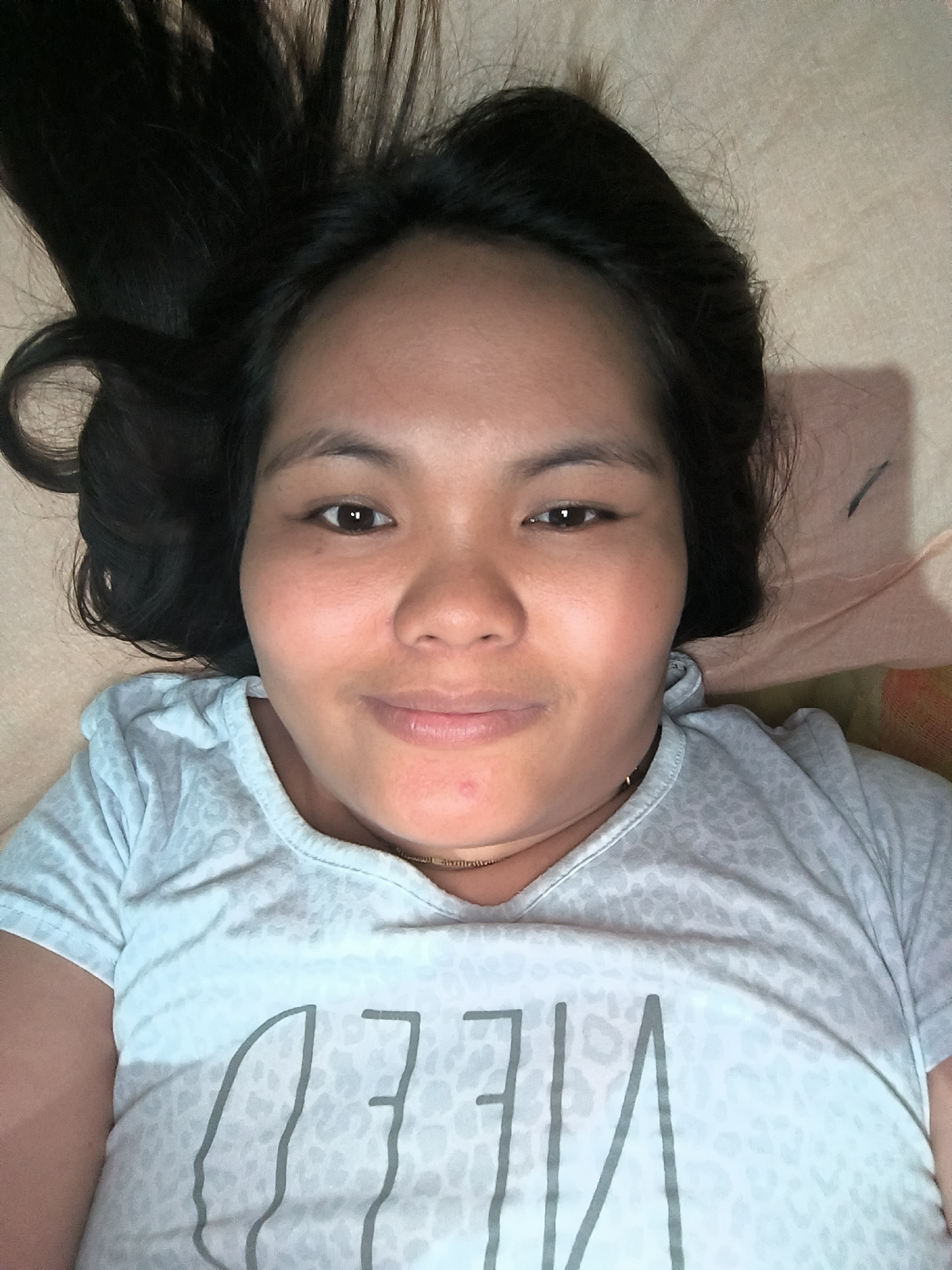 A woman with dark hair is lying down on a beige surface, possibly a bed. She is wearing a light-colored t-shirt with grey text. Her eyes are open and she is looking directly at the camera with a slight smile. She is also wearing a necklace. The location appears to be indoors.