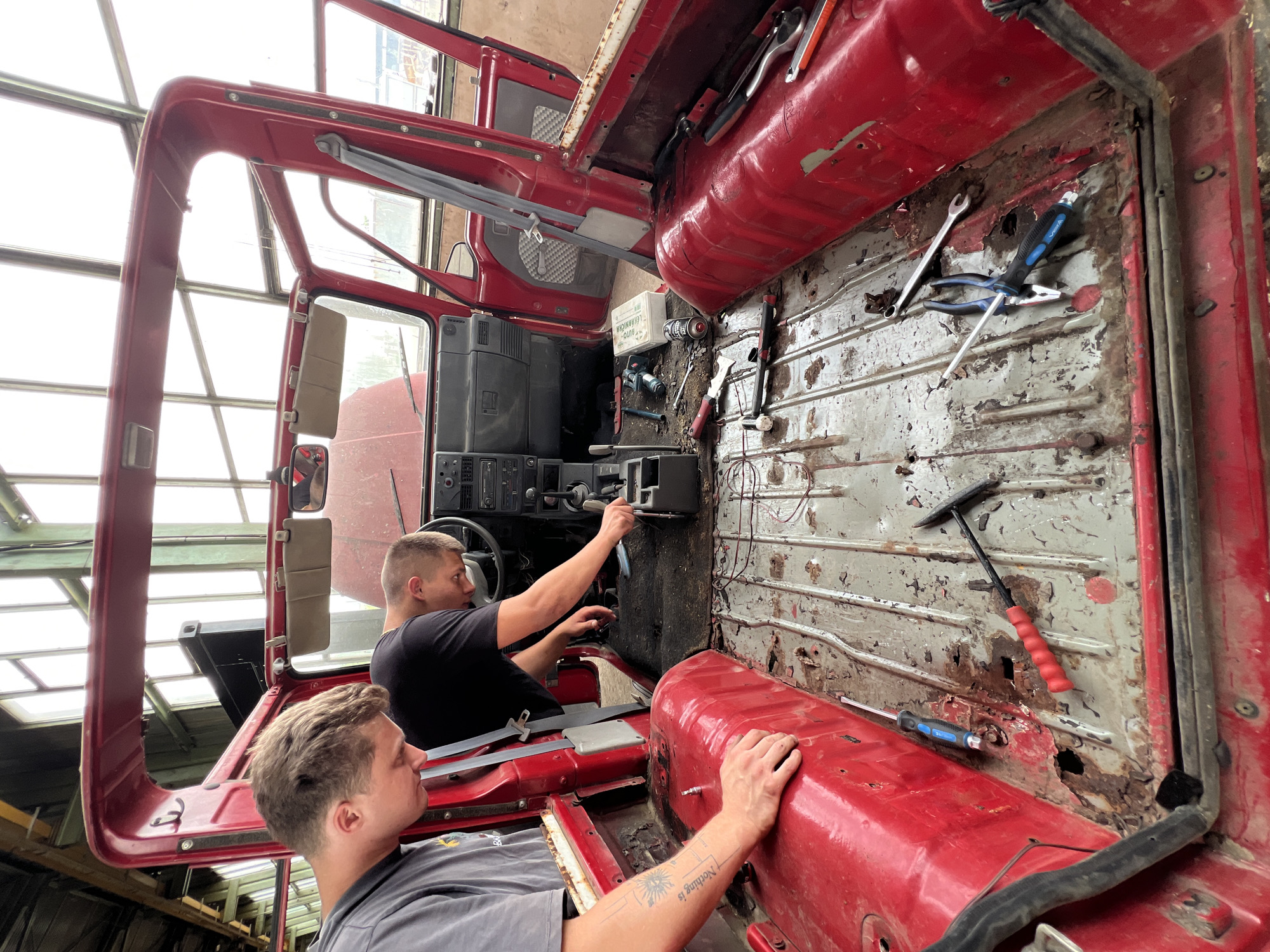 Two individuals are working inside the disassembled interior of a red vehicle, possibly undergoing repair or restoration. The vehicle's doors are open, and the roof is off, exposing the interior to natural light from a large, glass-paneled industrial building. The floor of the vehicle is heavily rusted and appears to have been stripped of its original flooring. Tools such as screwdrivers, pliers, a hammer, and a wrench are scattered across the floor. One individual, wearing a gray t-shirt, is seen with their hands on the red interior paneling of the vehicle. The other individual, wearing a black t-shirt, is leaning over the center console, appearing to work on the gear shift or dashboard area. The environment suggests a workshop or garage setting within Lanžhot, Czechia, characterized by industrial architecture and diffused natural light.