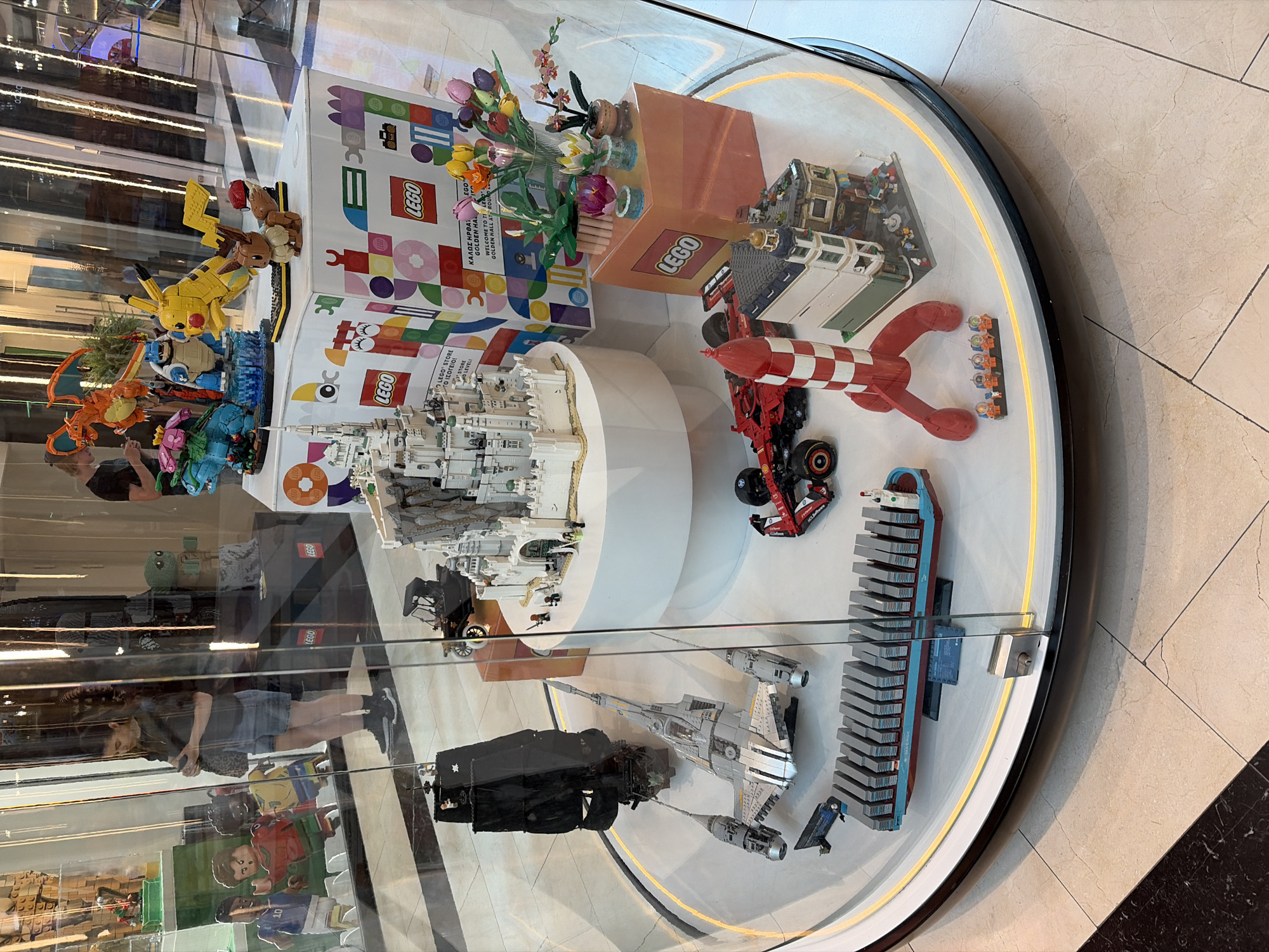 The image shows a circular display case filled with LEGO models. The display is located in a commercial area, likely a shopping mall, as evidenced by the polished floors and the presence of other shops in the background.
Inside the display case, various LEGO creations are arranged on a white circular platform. Prominent among the models are:
* A detailed replica of a medieval castle.
* A black pirate ship with tattered sails.
* A futuristic gray spaceship.
* A red and white striped rocket.
* A red Formula 1 style race car.
* A model of a cargo ship filled with containers.
* Several LEGO buildings, including a multi-story structure with detailed facades.
* A collection of LEGO Pokémon figures, including Pikachu, Charizard, Blastoise, and Venusaur.
* A bouquet of colorful LEGO flowers in vases.
In the background, behind the display case, two people are visible. One is a woman in a dark shirt and denim shorts, looking down at her phone. The other is a man standing partially obscured. The backdrop consists of shop interiors with shelves and displays, suggesting a retail environment. The lighting is bright, illuminating the LEGO models within the case. The display case is made of glass and appears to be a standalone exhibit.