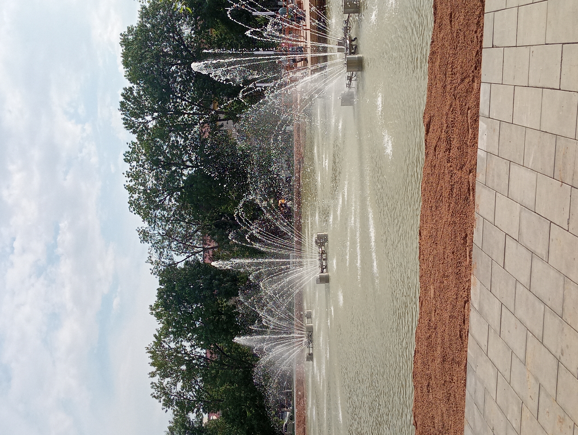 The image shows a public park in Shymkent, Kazakhstan, featuring a large pond with multiple active fountains spraying water upwards. The pond is surrounded by a decorative border of reddish-brown mulch and a paved walkway made of light gray square tiles in the foreground. In the background, lush green trees form a dense line, with glimpses of buildings and people visible between them on what appears to be a promenade or seating area. The sky is partly cloudy with a mix of white and gray clouds against a light blue background. The overall scene depicts a serene outdoor environment designed for recreation and relaxation.
